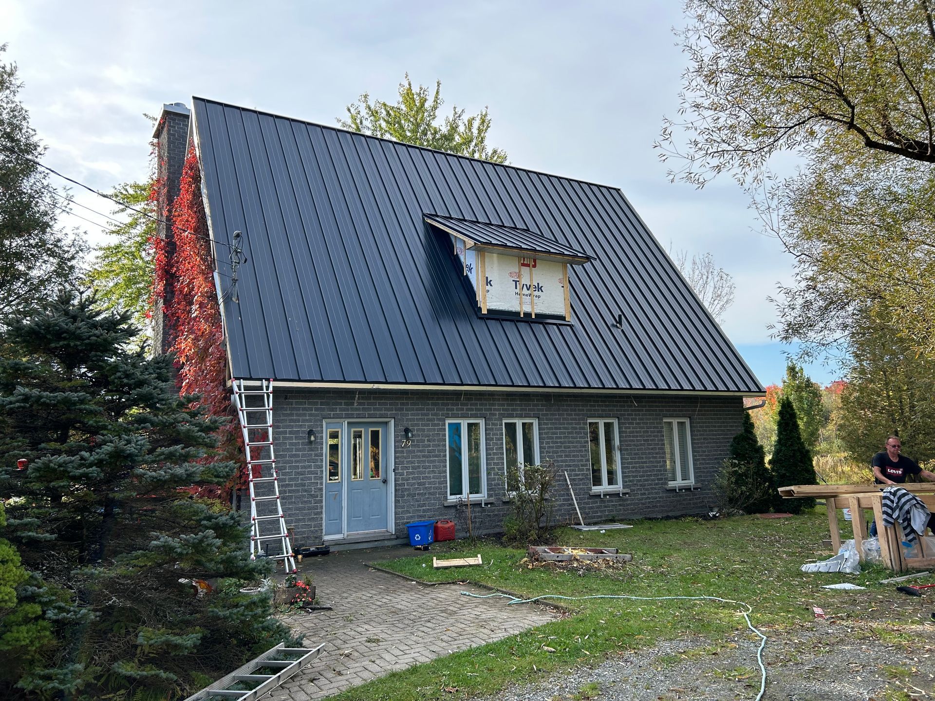 Extérieur de la maison avec bardage gris foncé et nouvelle toiture en métal noir, une haute cheminée en briques recouverte de vigne rouge.