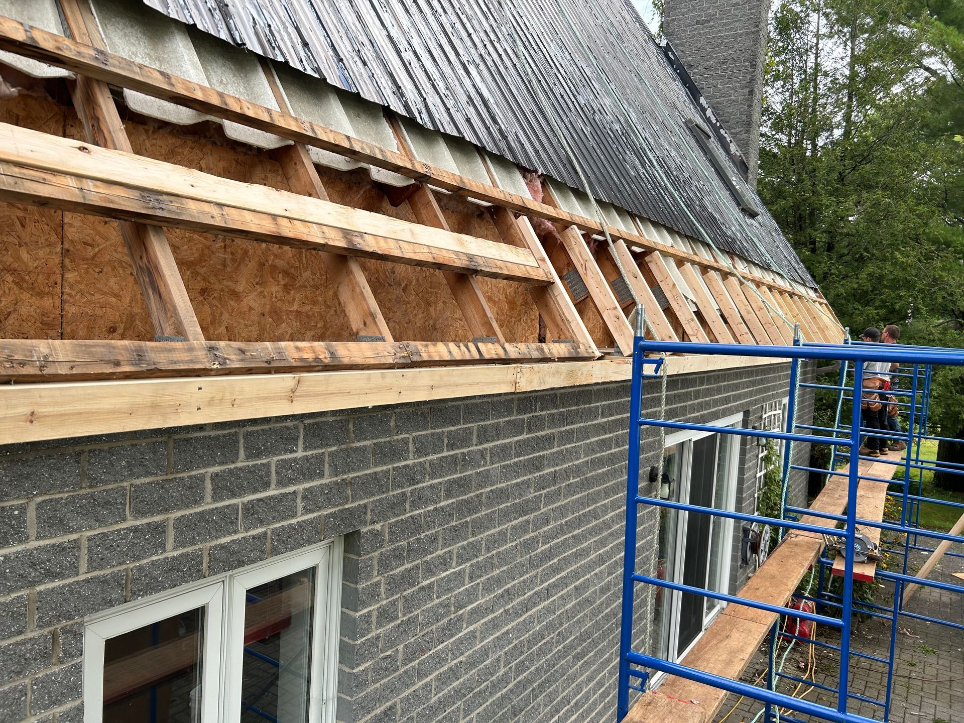 Travaux de construction sur le toit d'un bâtiment en briques avec poutres en bois apparentes, échafaudages et matériaux de couverture partiellement installés.