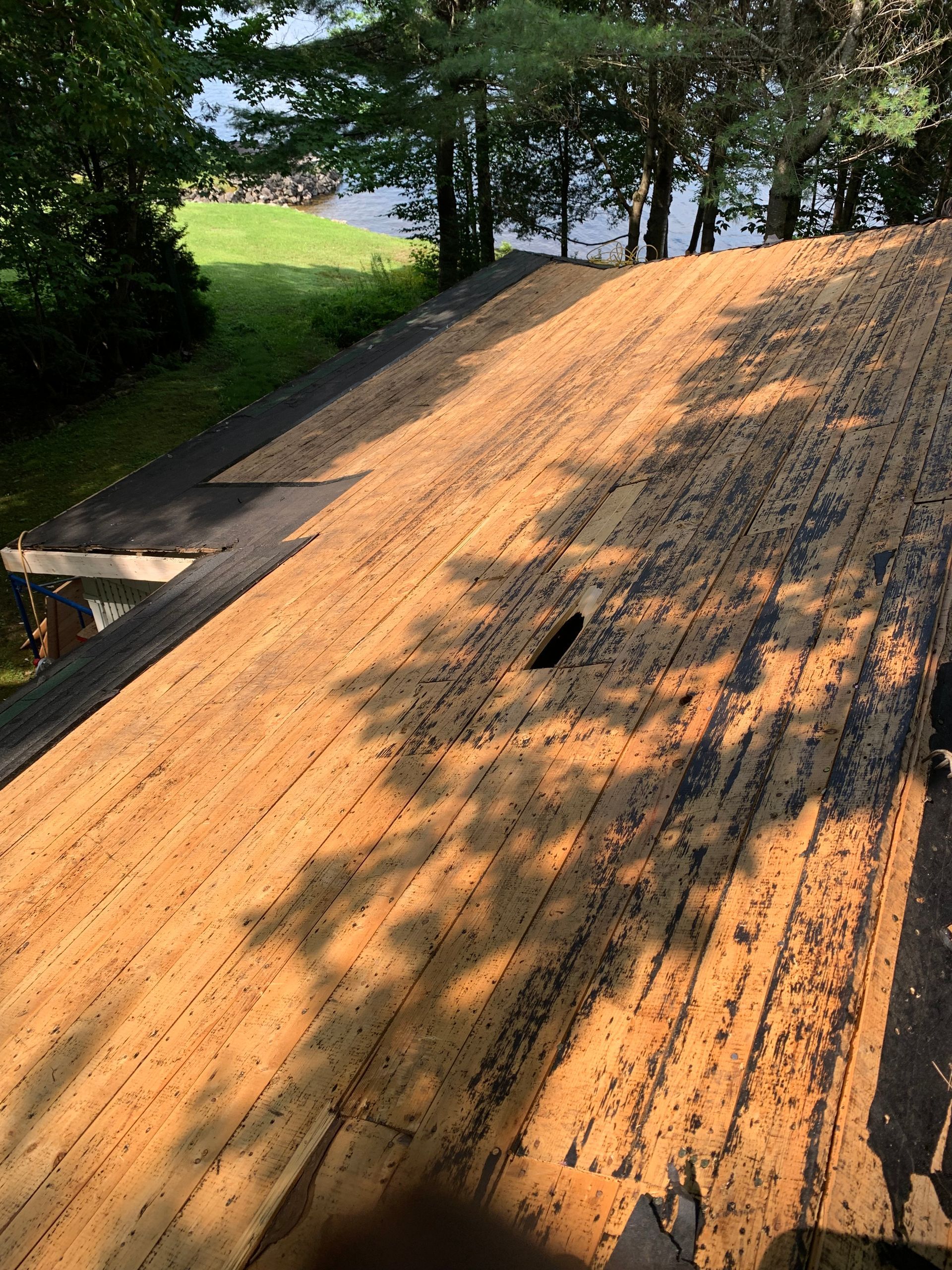 Toiture dont les tuiles ont été retirées, laissant apparaître la sous-couche. À l'extérieur, arbres et eau en arrière-plan.