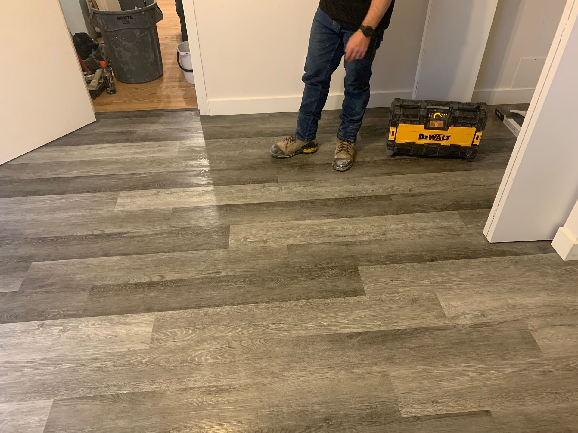Une personne se tient debout sur un revêtement de sol gris imitation bois, une boîte à outils se trouvant à proximité.
