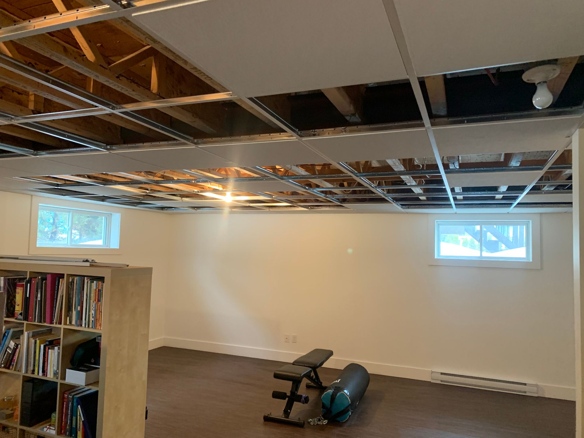Rénovation du sous-sol en cours, plafond partiellement enlevé. Murs blancs, plancher de bois, banc de musculation. Deux fenêtres.