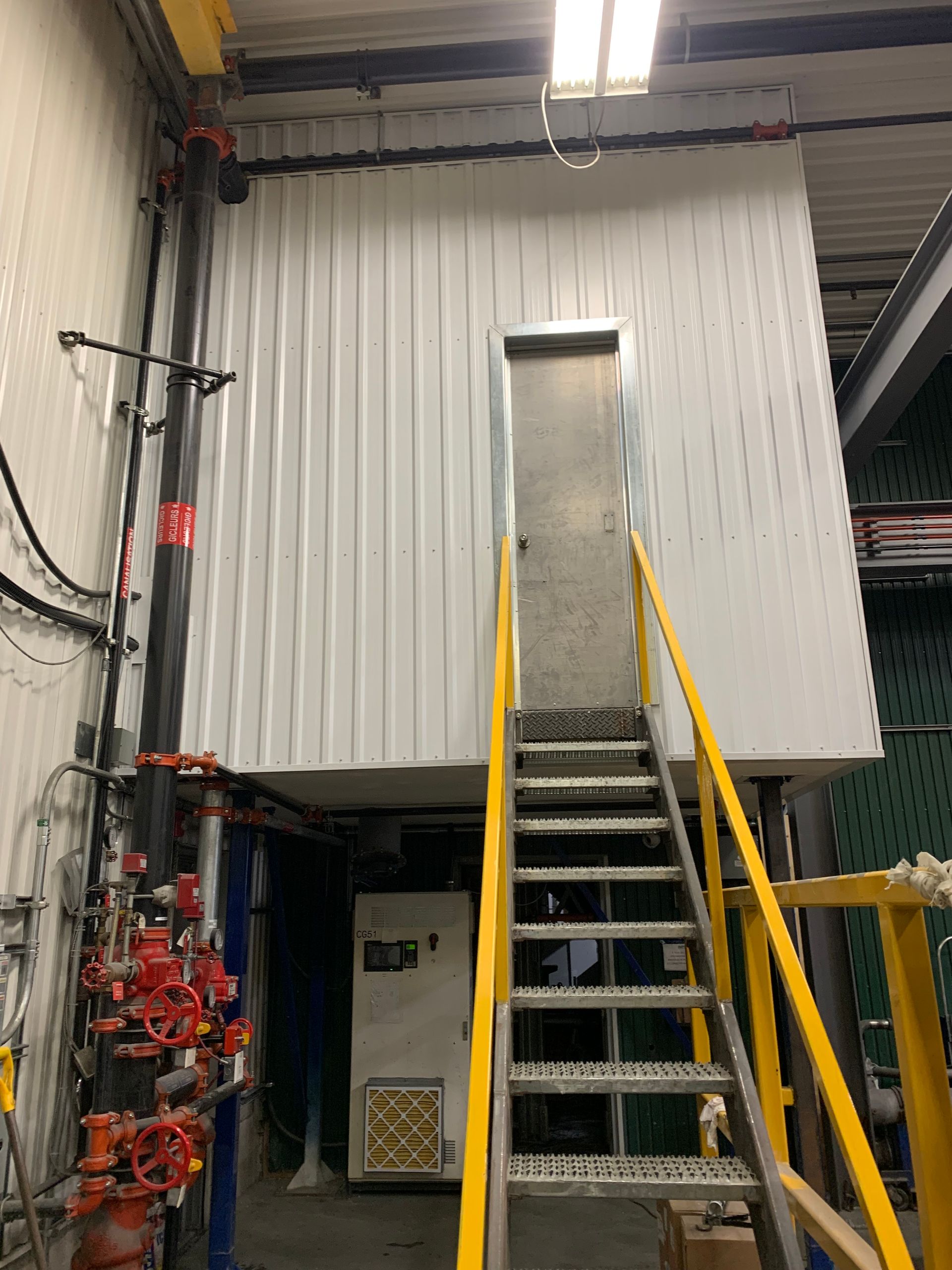 Un escalier métallique mène à une plateforme munie d'une porte, dans un environnement industriel.