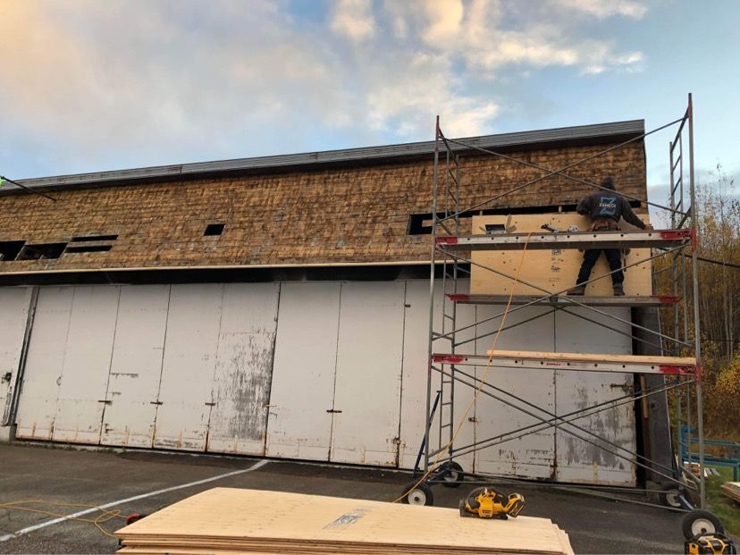 Une personne sur un échafaudage installe du contreplaqué sur le toit d'un bâtiment recouvert de vieux bardeaux de bois.