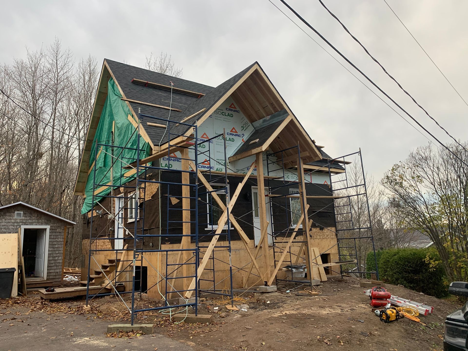 Maison en construction ; extérieur avec charpente en bois apparente, échafaudages et bâche verte.