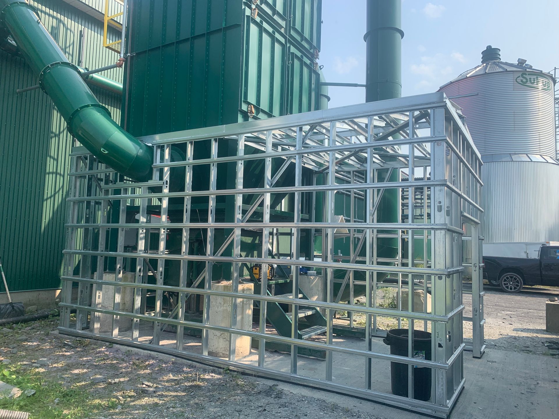 Cage de sécurité métallique autour d'un équipement industriel, avec un tuyau vert et un silo en arrière-plan.