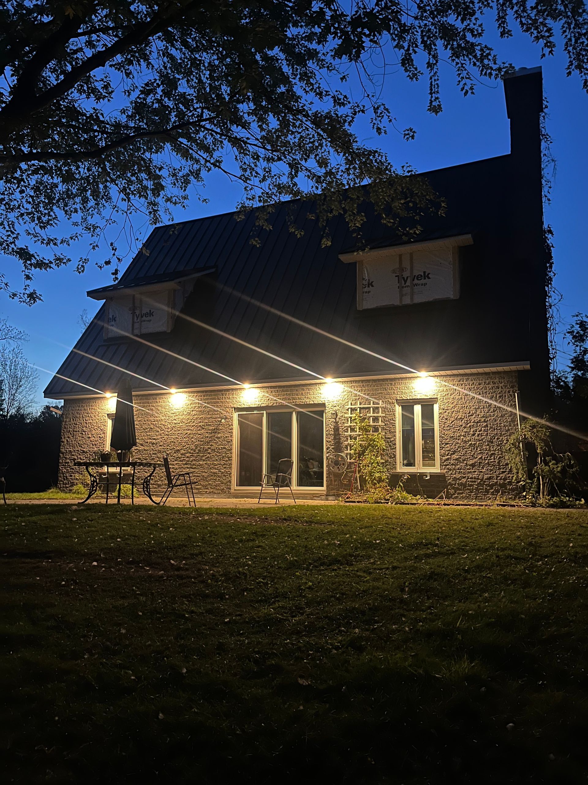 Maison en pierre illuminée au crépuscule, avec porche éclairé, cheminée et ciel sombre.