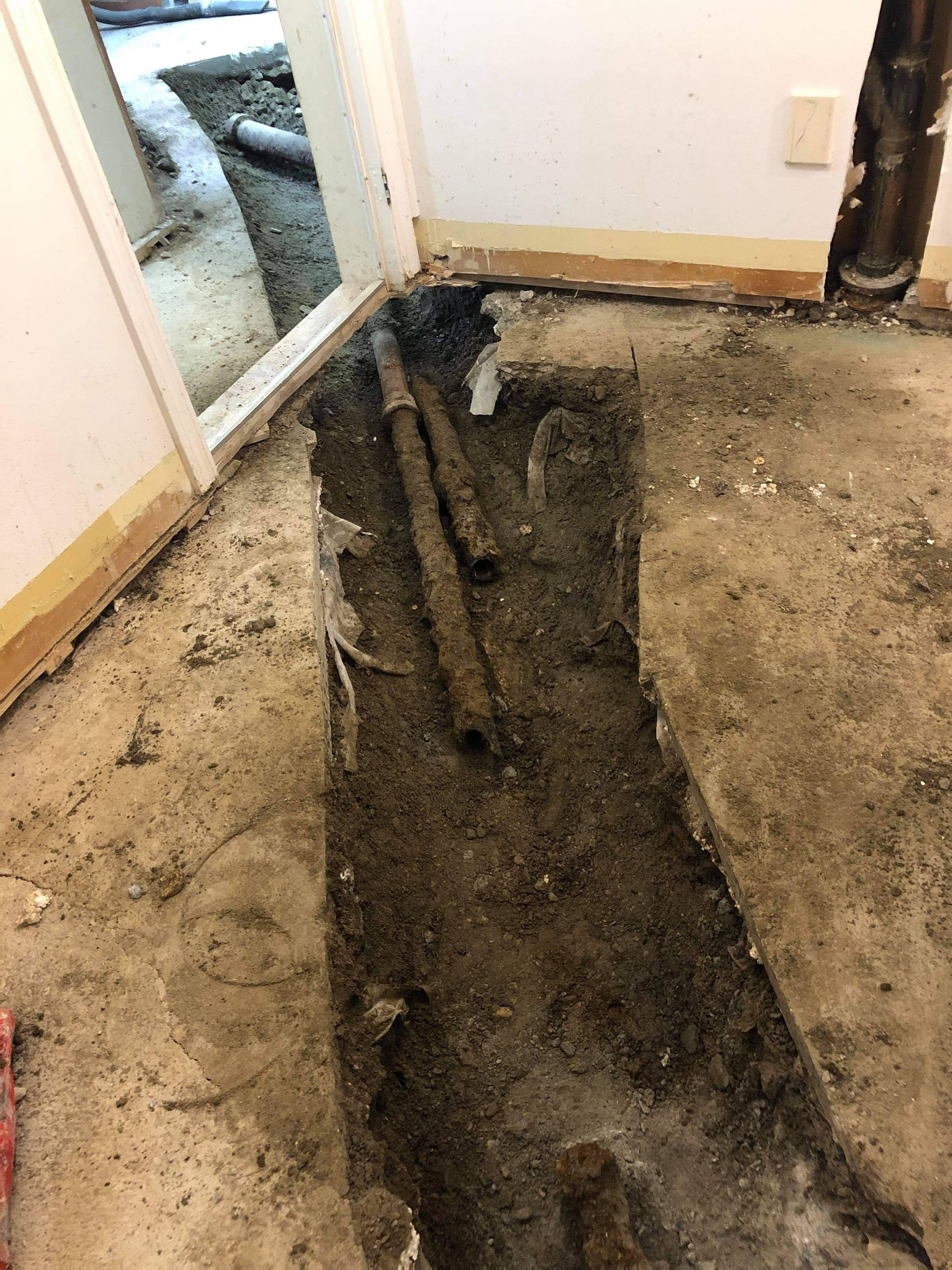 Tranchée ouverte dans le sol, laissant apparaître des canalisations. Travaux de rénovation en cours près d'une porte.