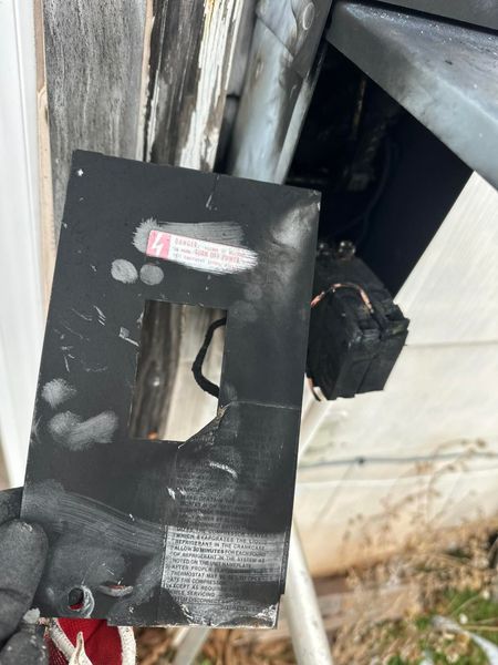 Damaged black electrical panel with a hole, held next to charred wires, likely from a fire.