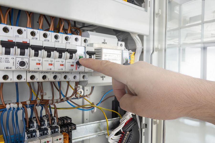 Hand pointing at electrical breaker panel with wires and switches inside a control cabinet
