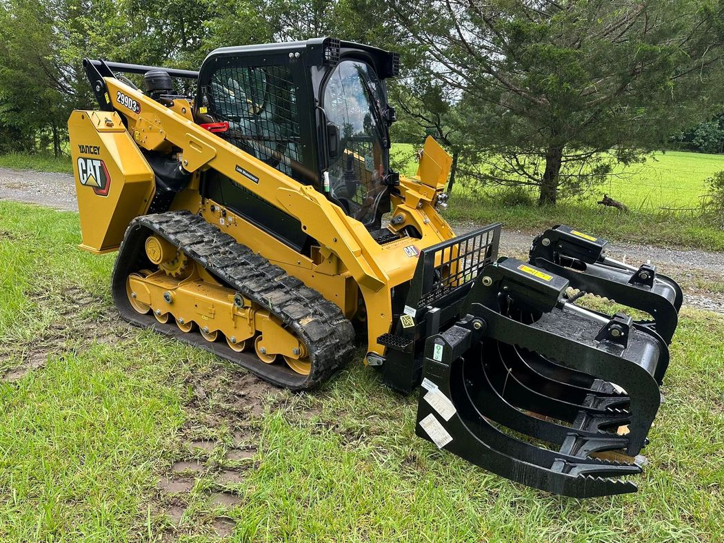 Yellow compact track loader with a front grapple attachment on grass near trees