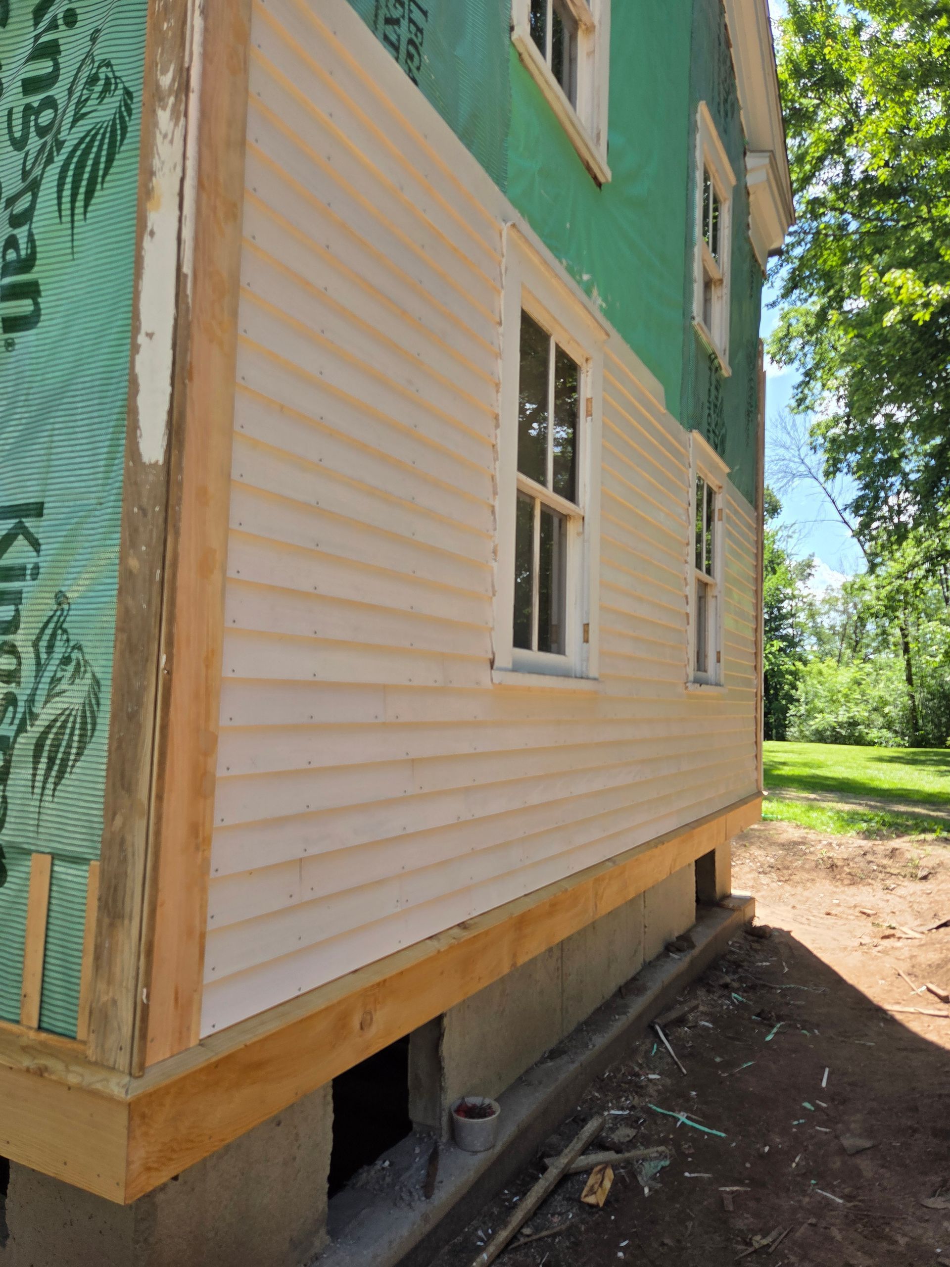 Side of a small house under construction with beige siding, green sheathing, and windows on a dirt lot.