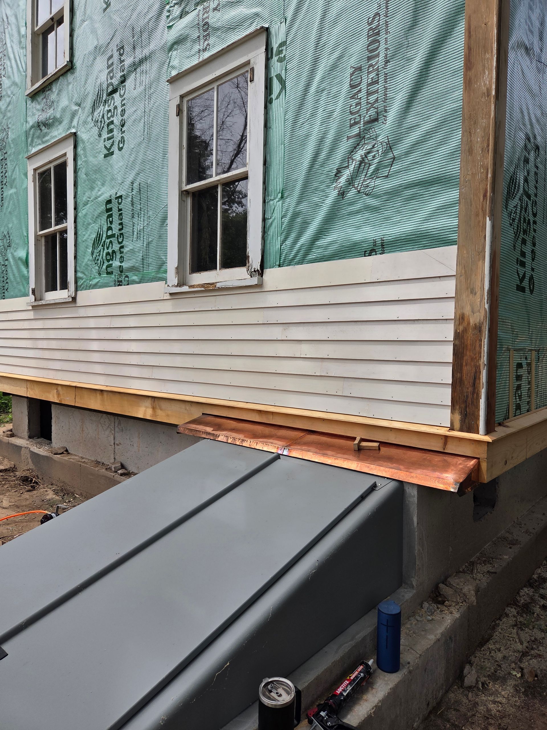 Partially renovated house exterior with green wrap, white siding, and a black metal awning over a basement entry