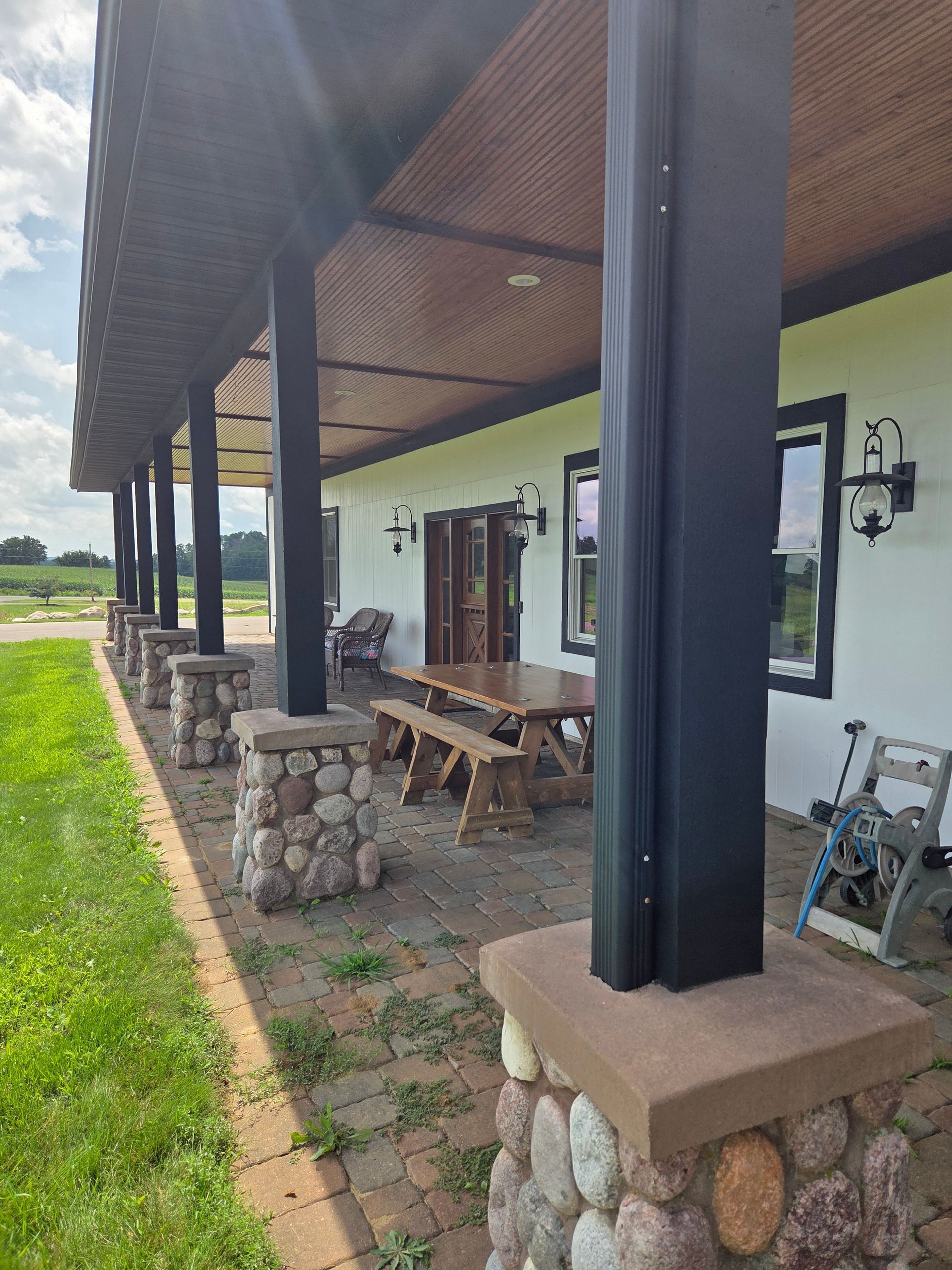 Covered outdoor patio with stone columns, picnic tables, and a grassy view beside a white building
