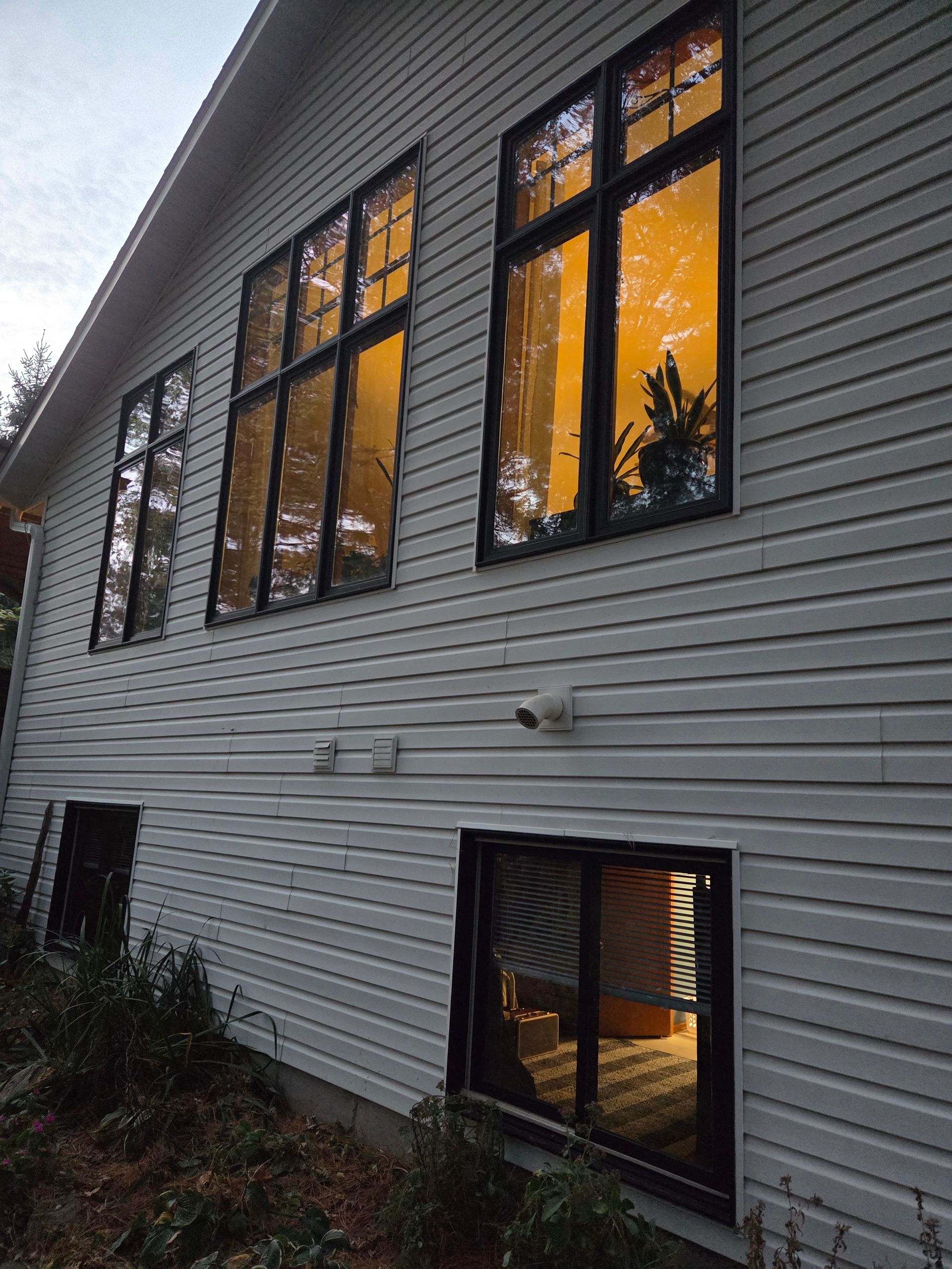 Side view of a gray house with lit windows glowing orange at dusk.