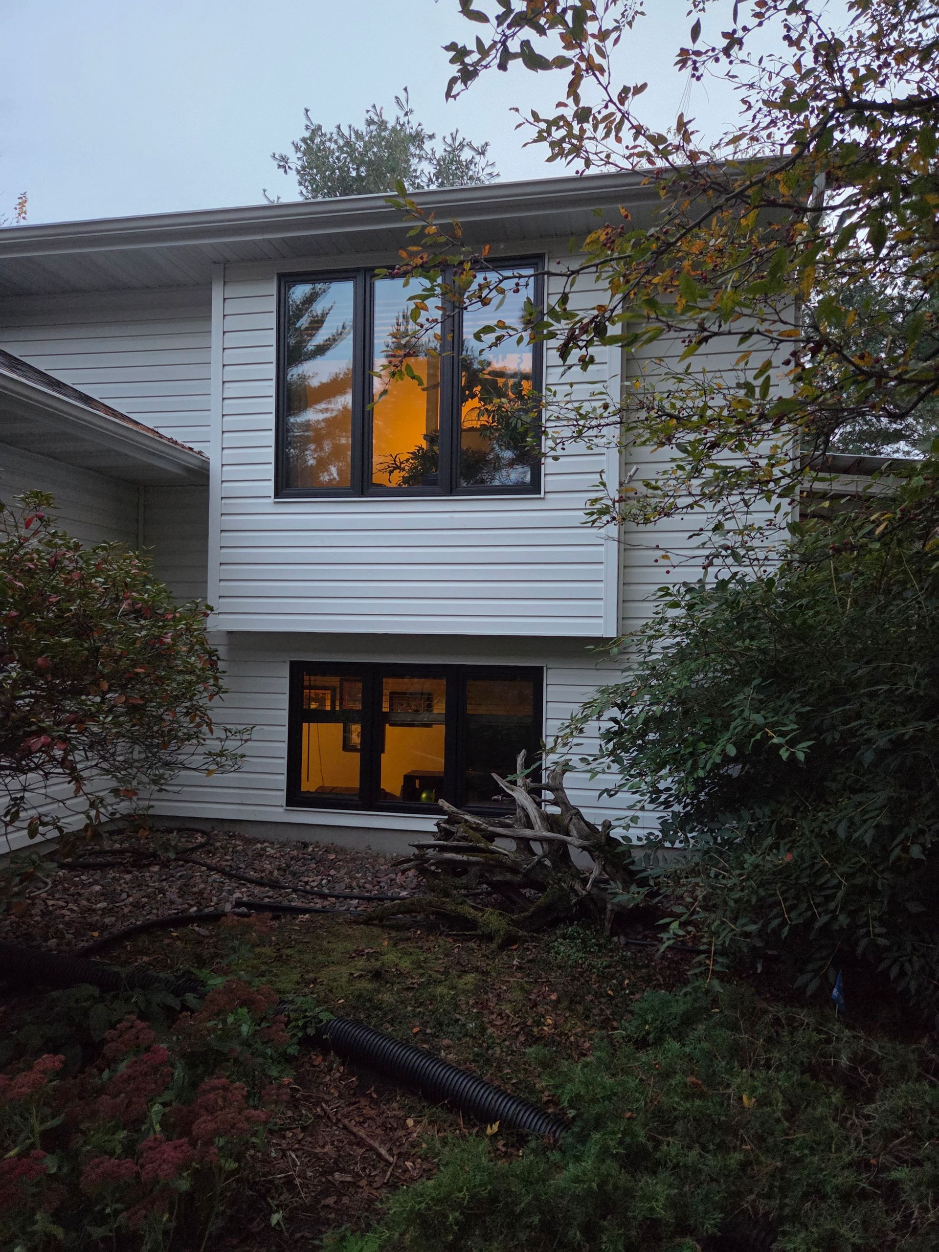Two-story white house with lit windows, seen through trees at dusk