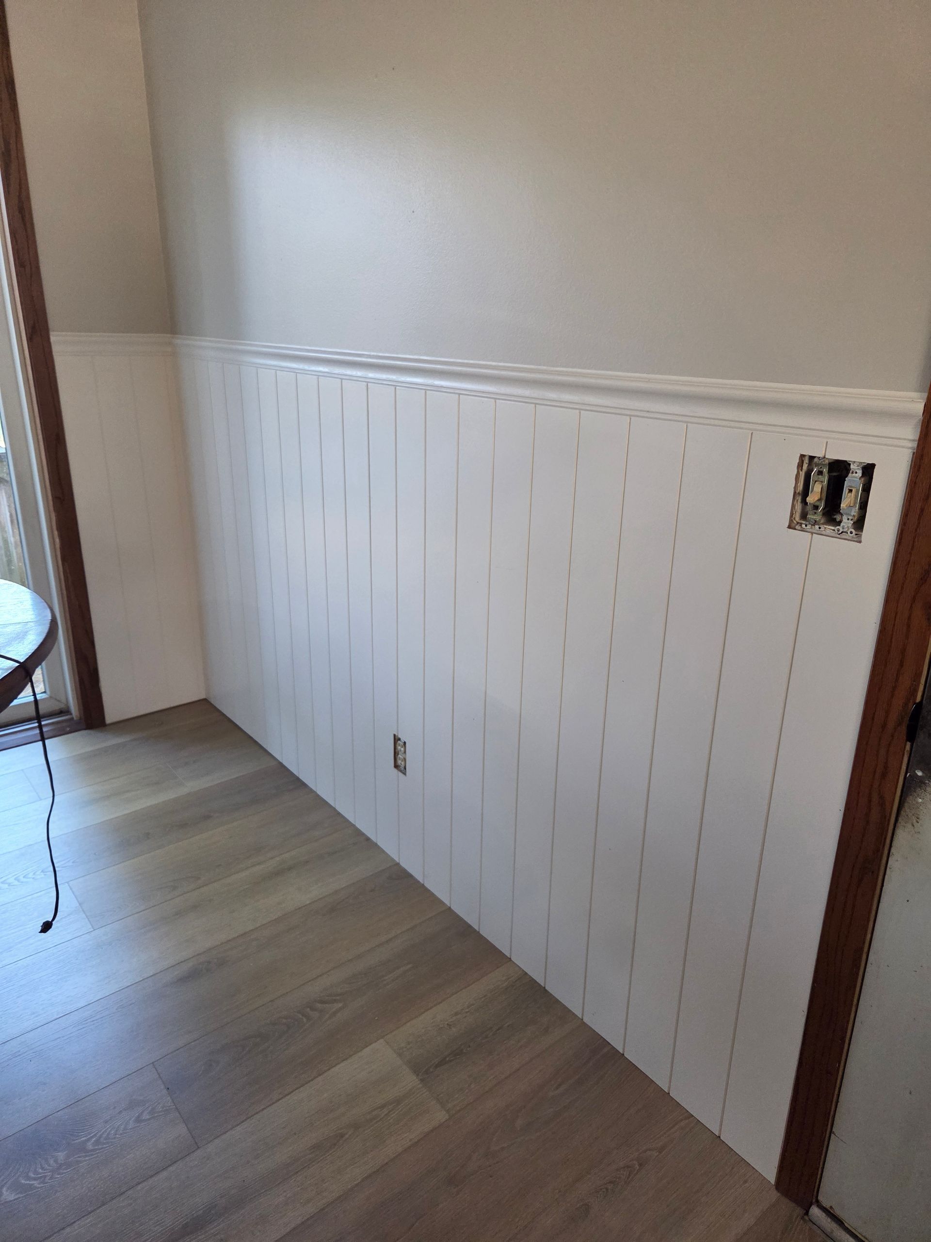 White beadboard wainscoting on a beige wall in a hallway with wood flooring and an electrical outlet cutout