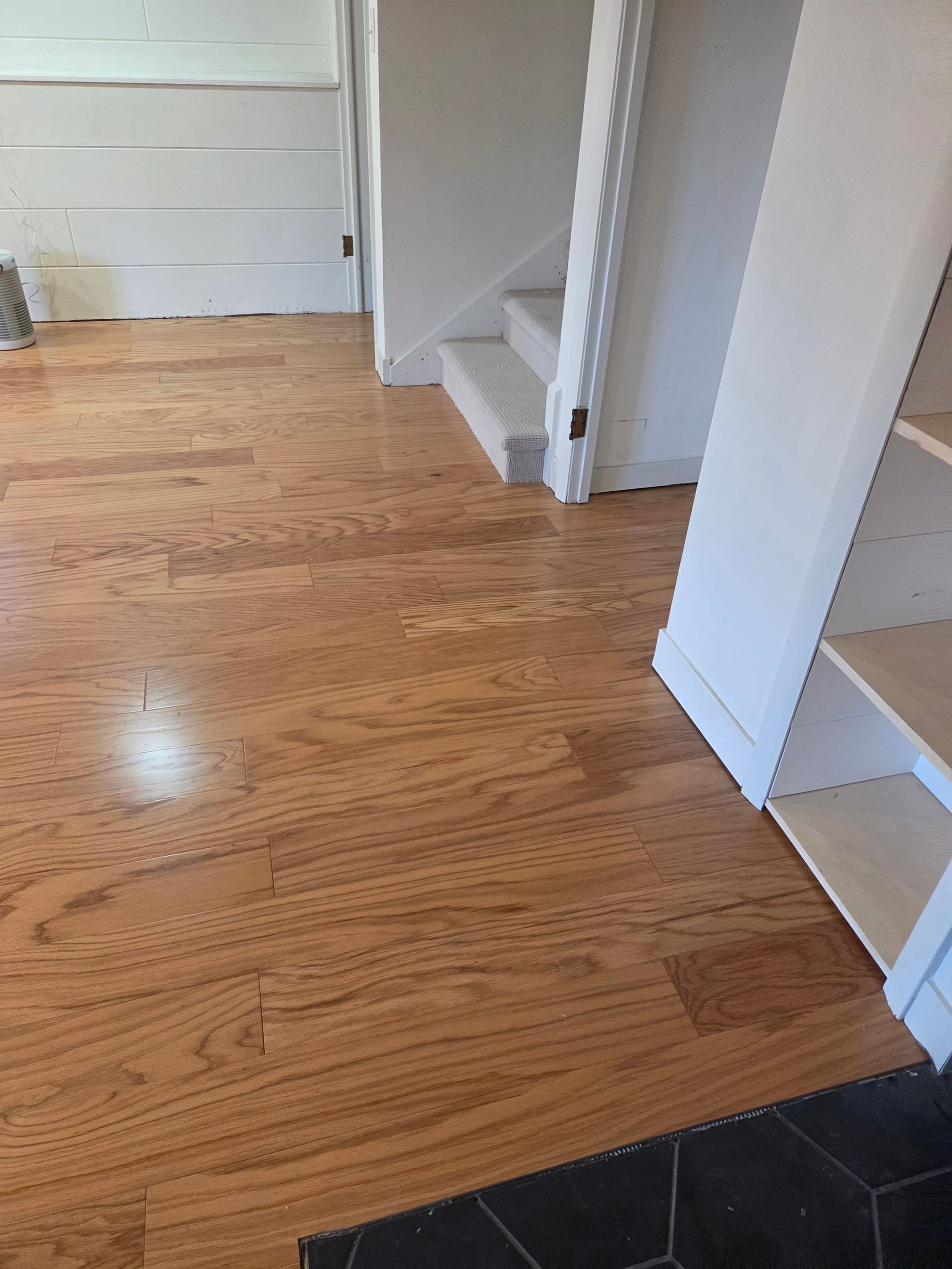 Wooden hallway floor with white walls, stairs, and a dark tile threshold