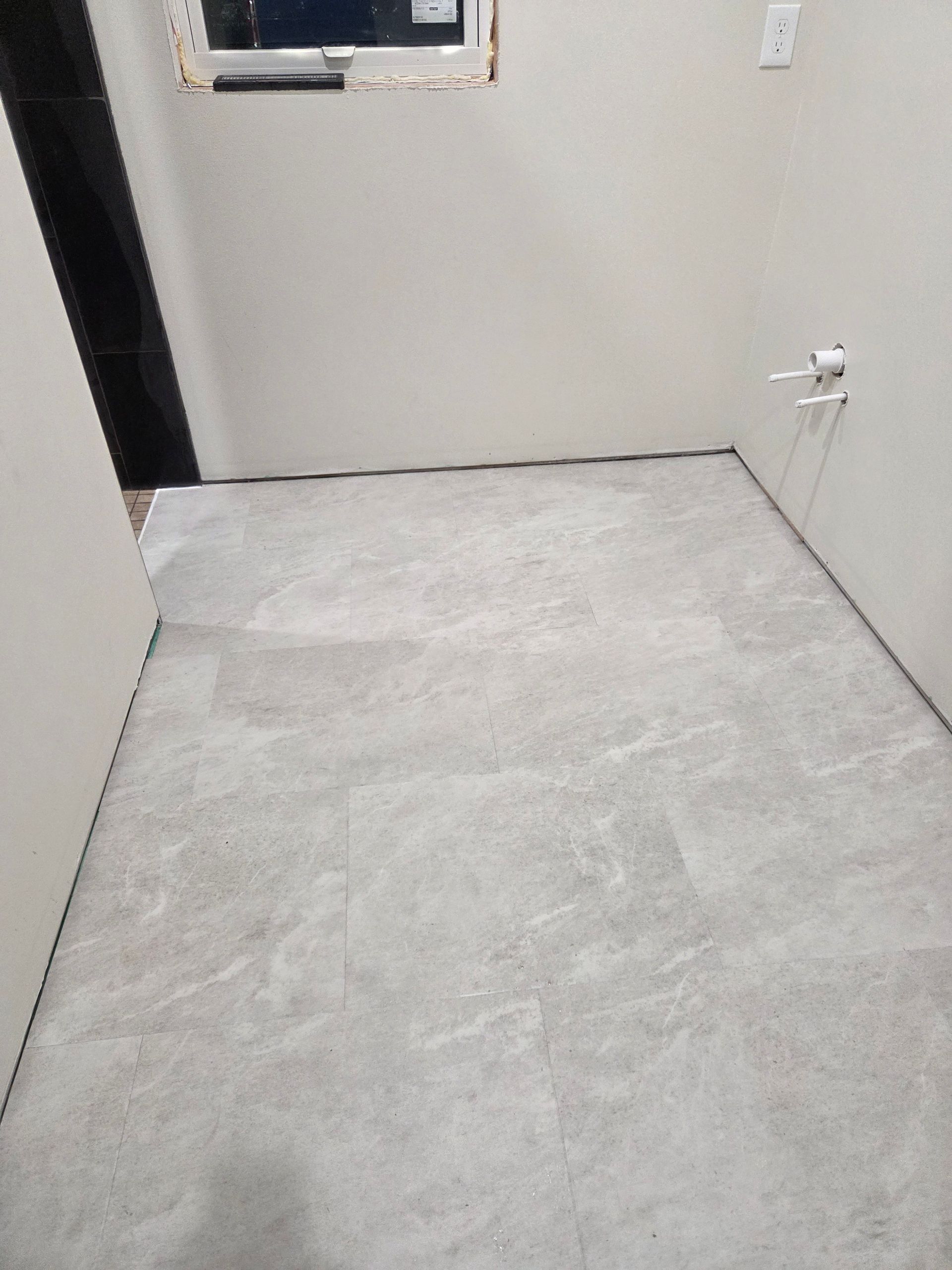 Empty room with light gray walls and floor, window opening, and electrical outlet on the right.