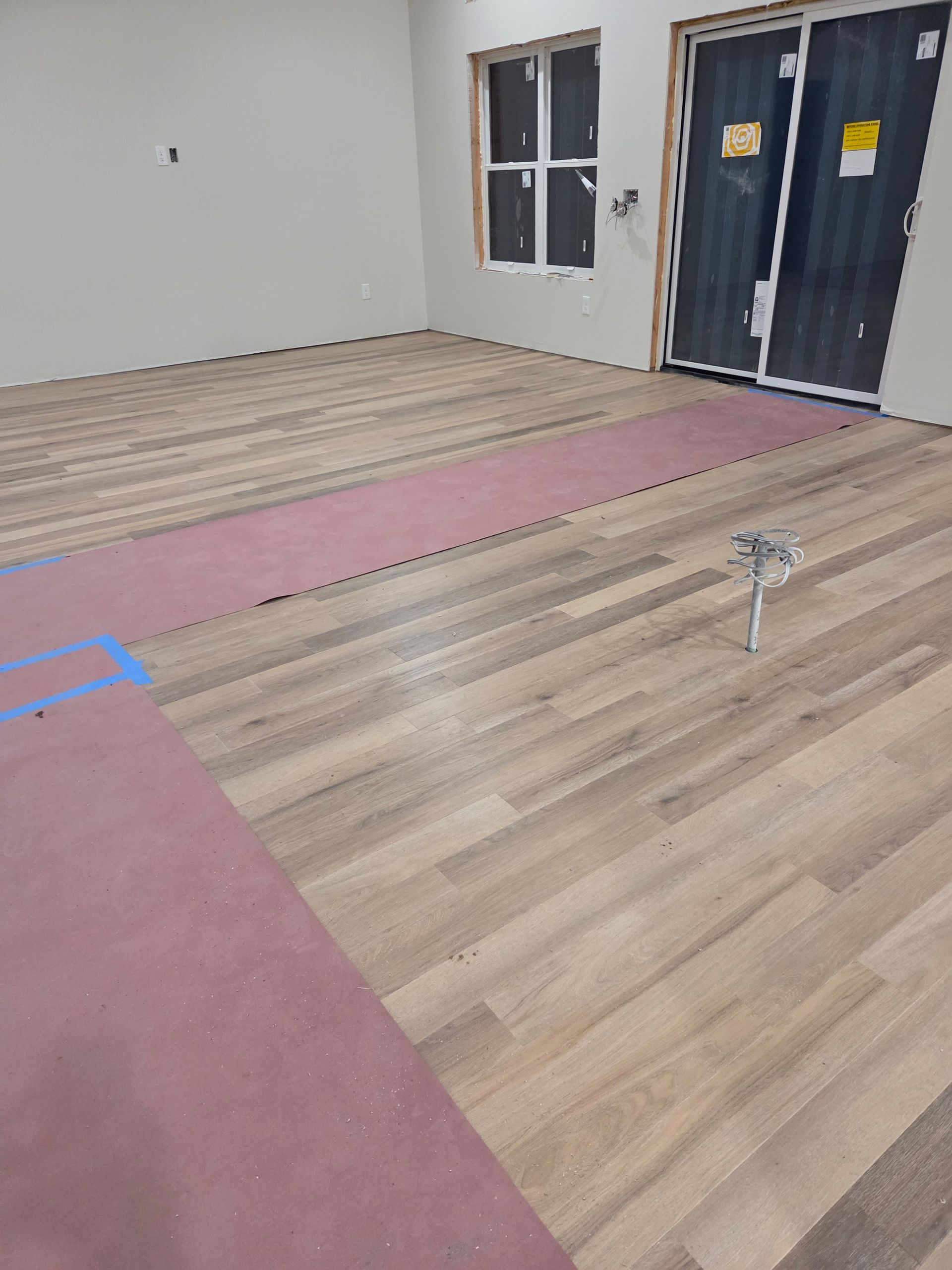 Empty room with wood flooring, pink underlayment, and windows near a black door.