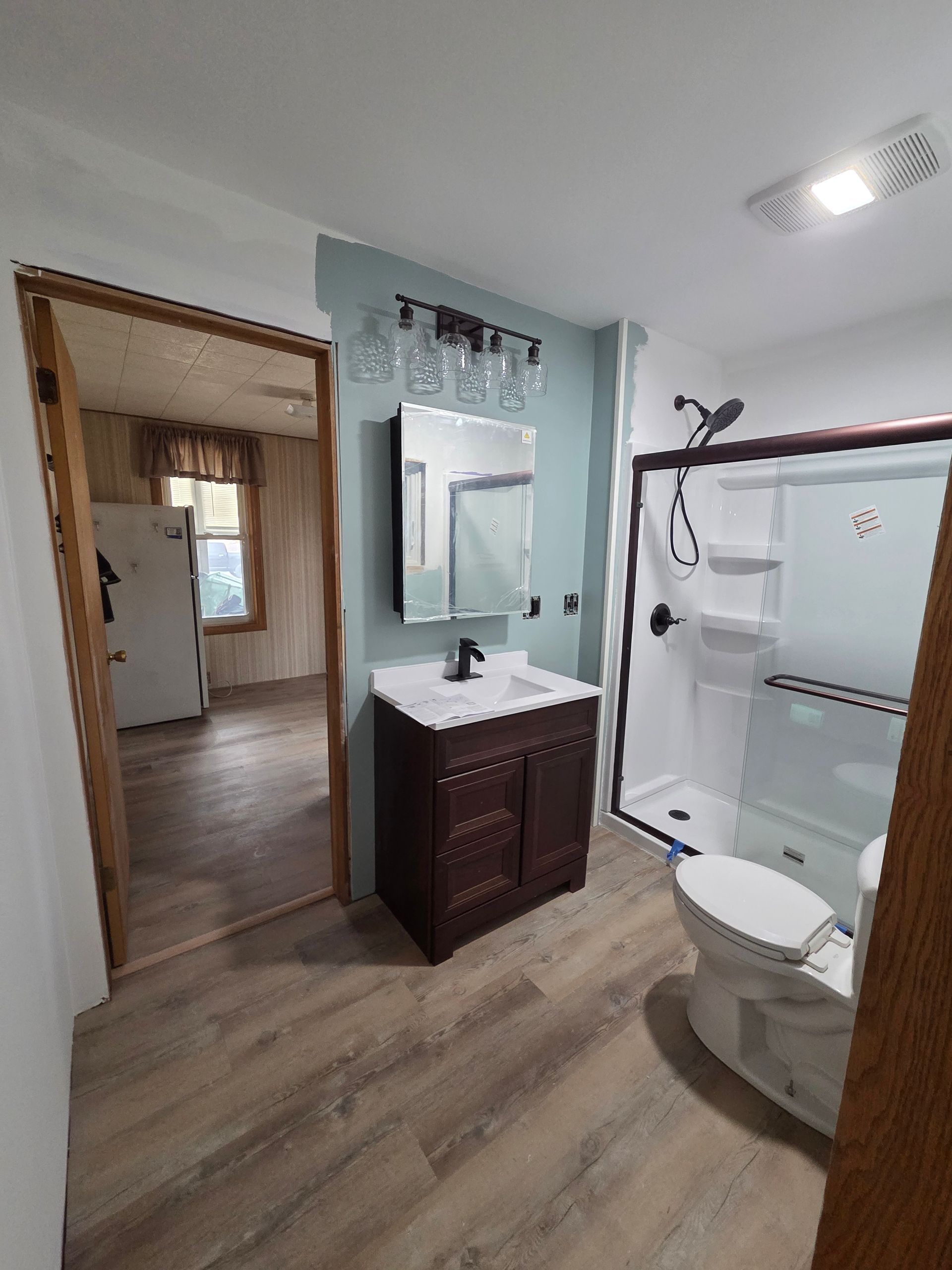 Small bathroom with white shower, toilet, dark vanity, mirror, and wood-look flooring; hallway visible through doorway