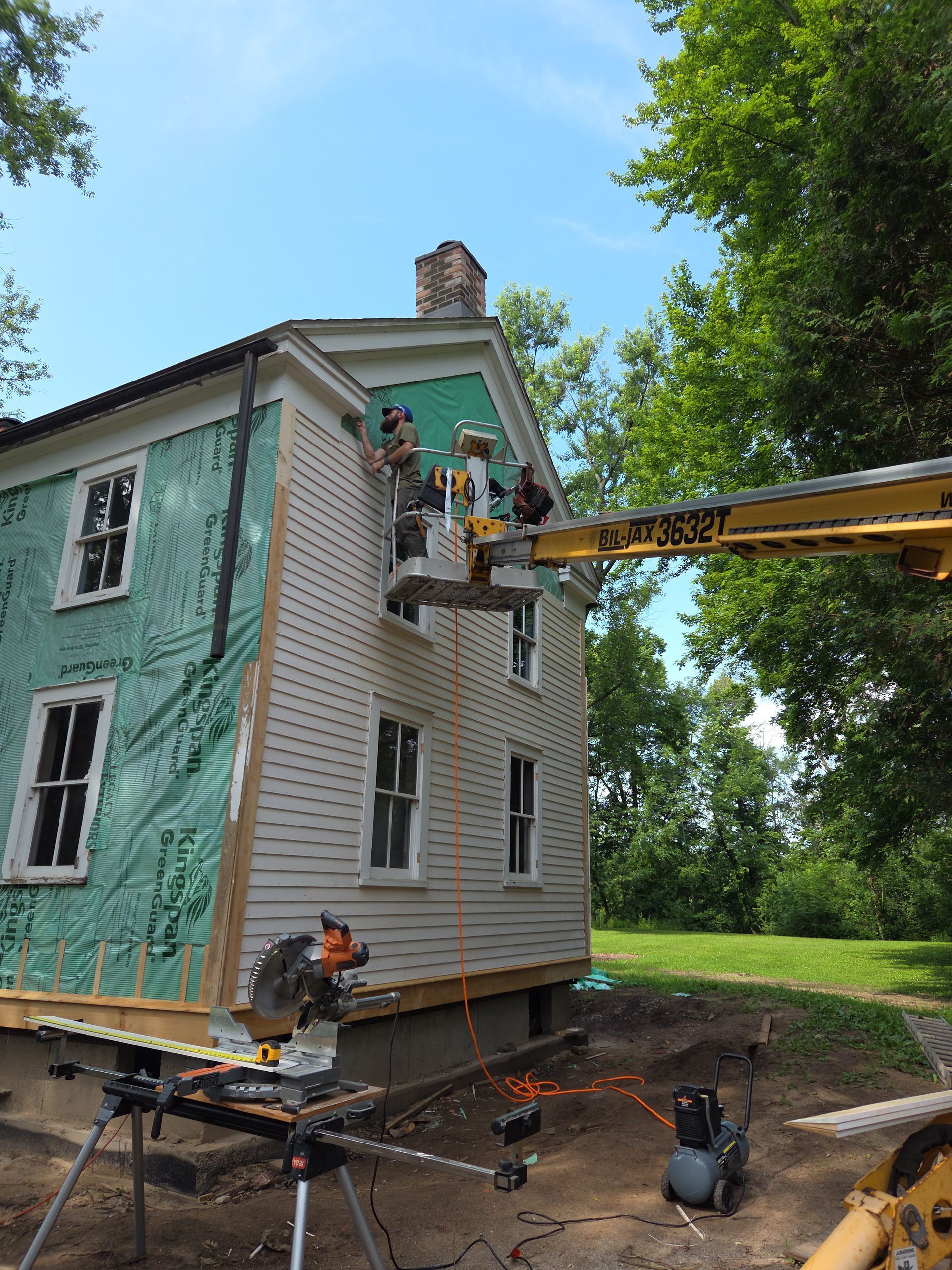 Workers painting a two-story house beside a lift bucket in a leafy yard