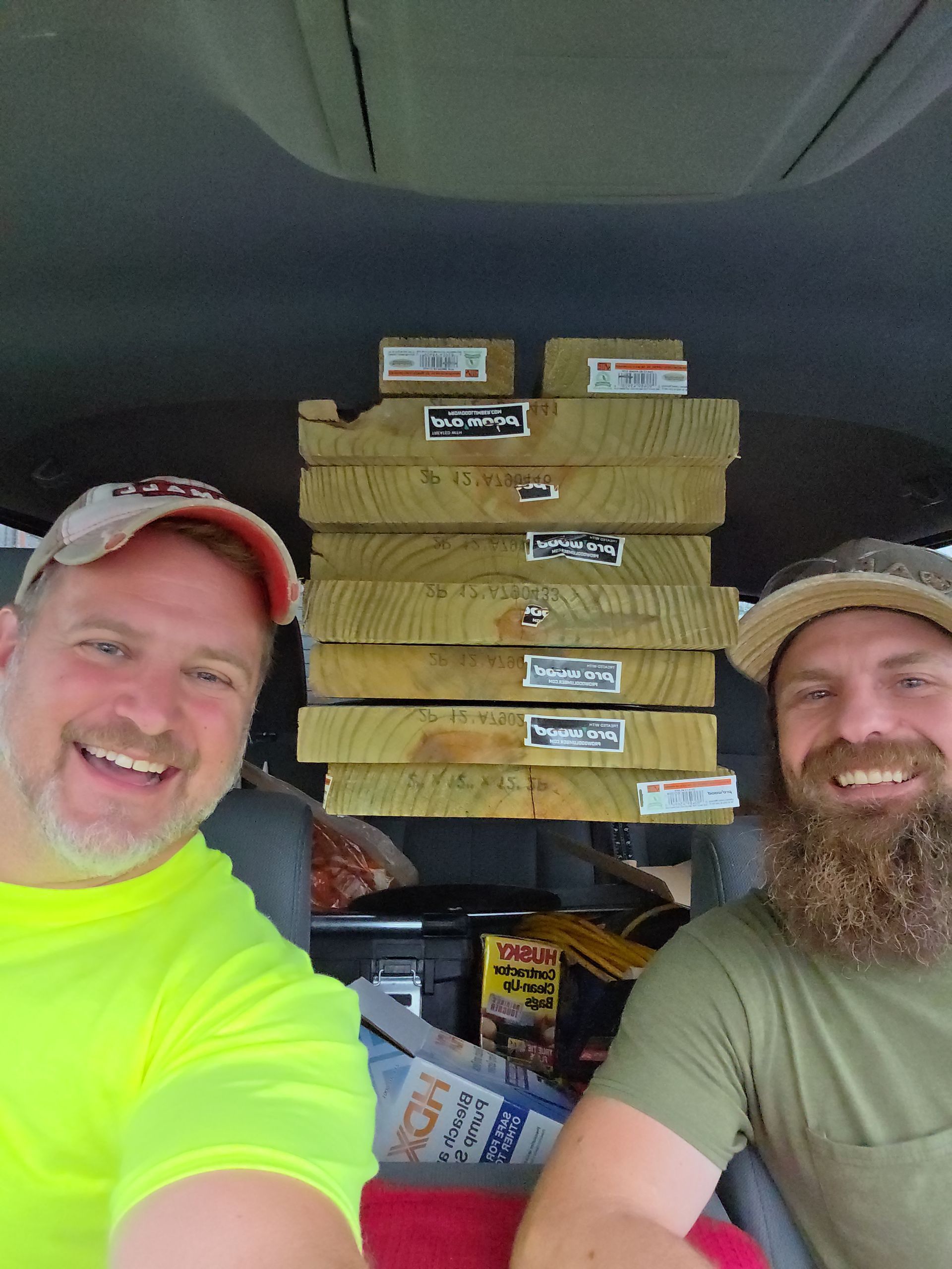 Two men smiling in a vehicle, with stacked pizza boxes between them.
