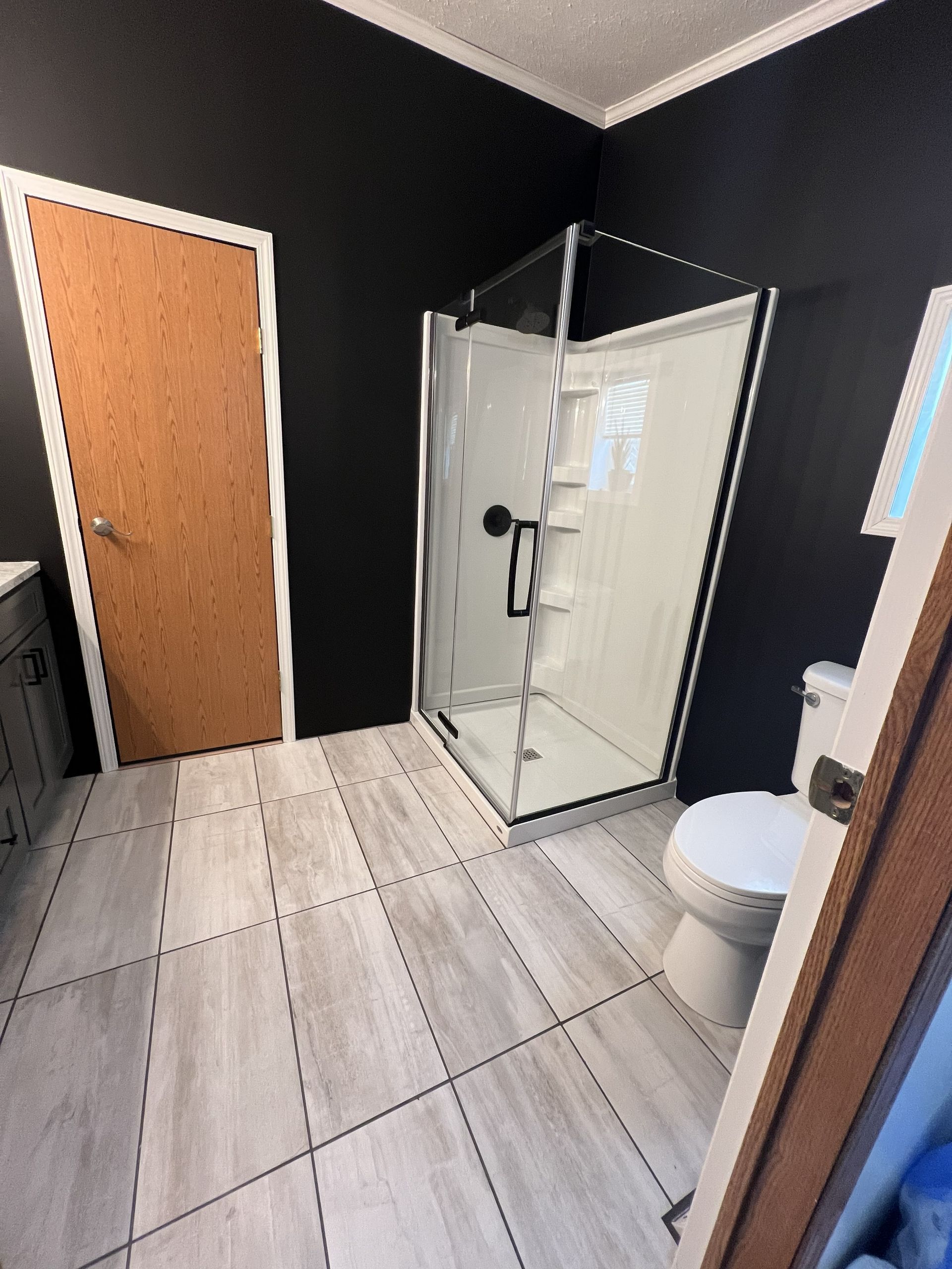 Small bathroom with black walls, white tiled floor, glass shower, toilet, and closed wooden door.