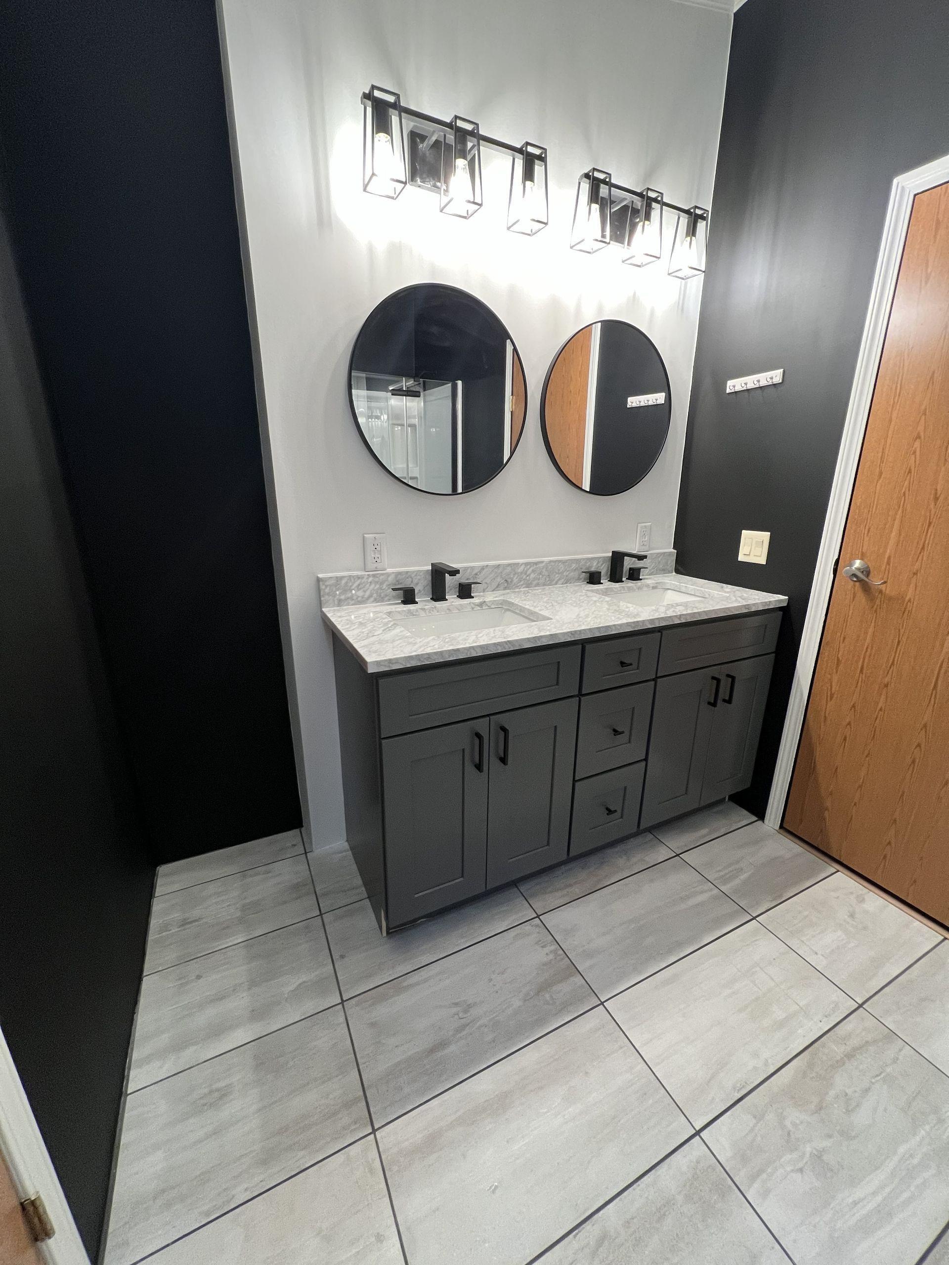 Modern restroom vanity with two round mirrors, gray cabinets, wall lights, and a wood door