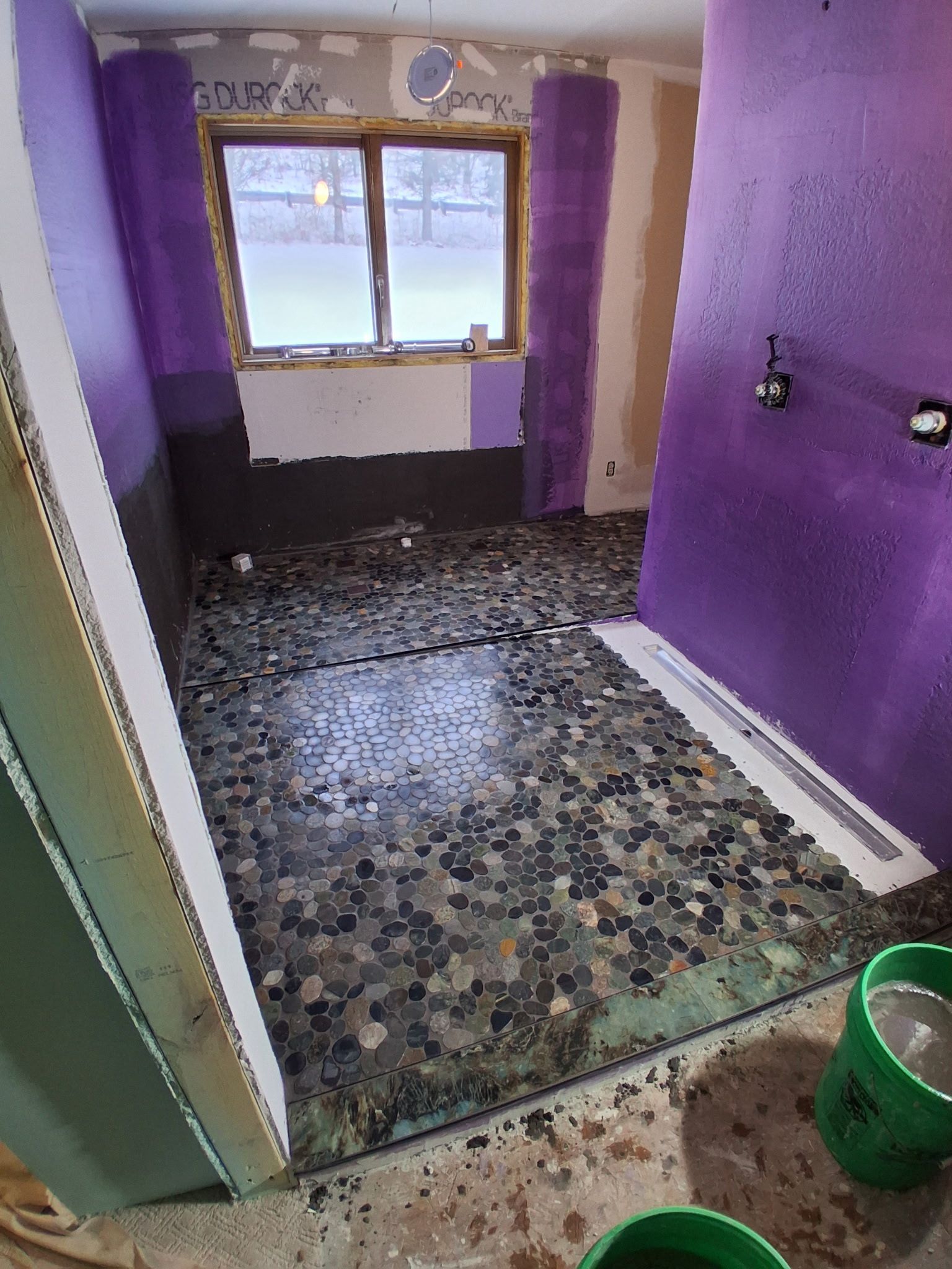 Small bathroom under renovation with purple walls, exposed floor, window, and a green bucket near the door.