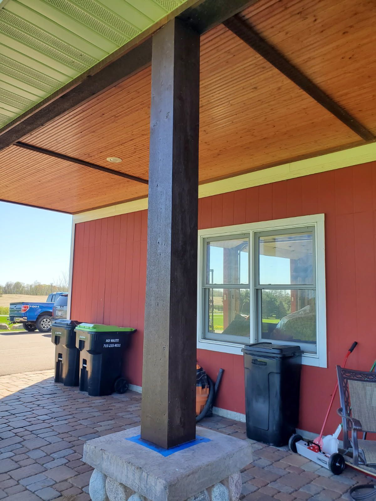 Covered porch with a dark support post, red wall, windows, and trash bins on a brick patio.