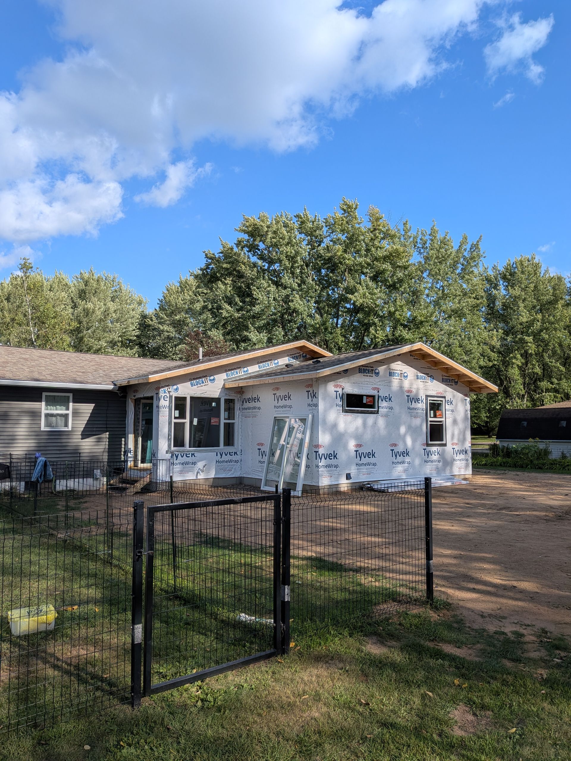 Partially renovated house with white exterior, black fence, and trees under a blue sky