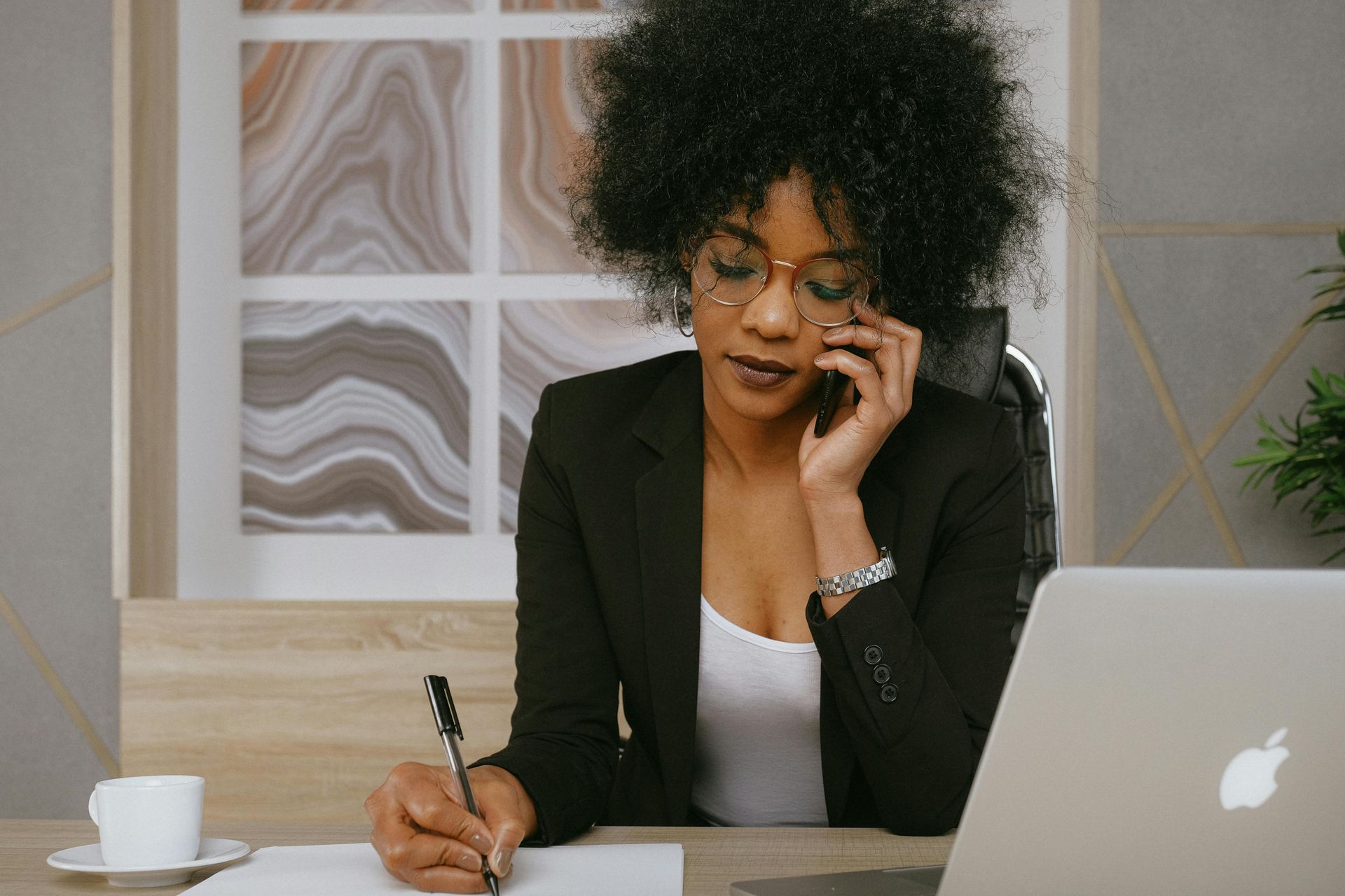 A woman is sitting at a desk talking on a cell phone and writing on a piece of paper.