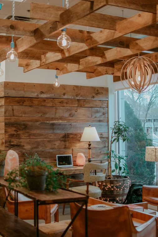 Cozy interior: wood-paneled walls, pallet ceiling, vintage lights, plants, laptop, and comfortable seating near a window.