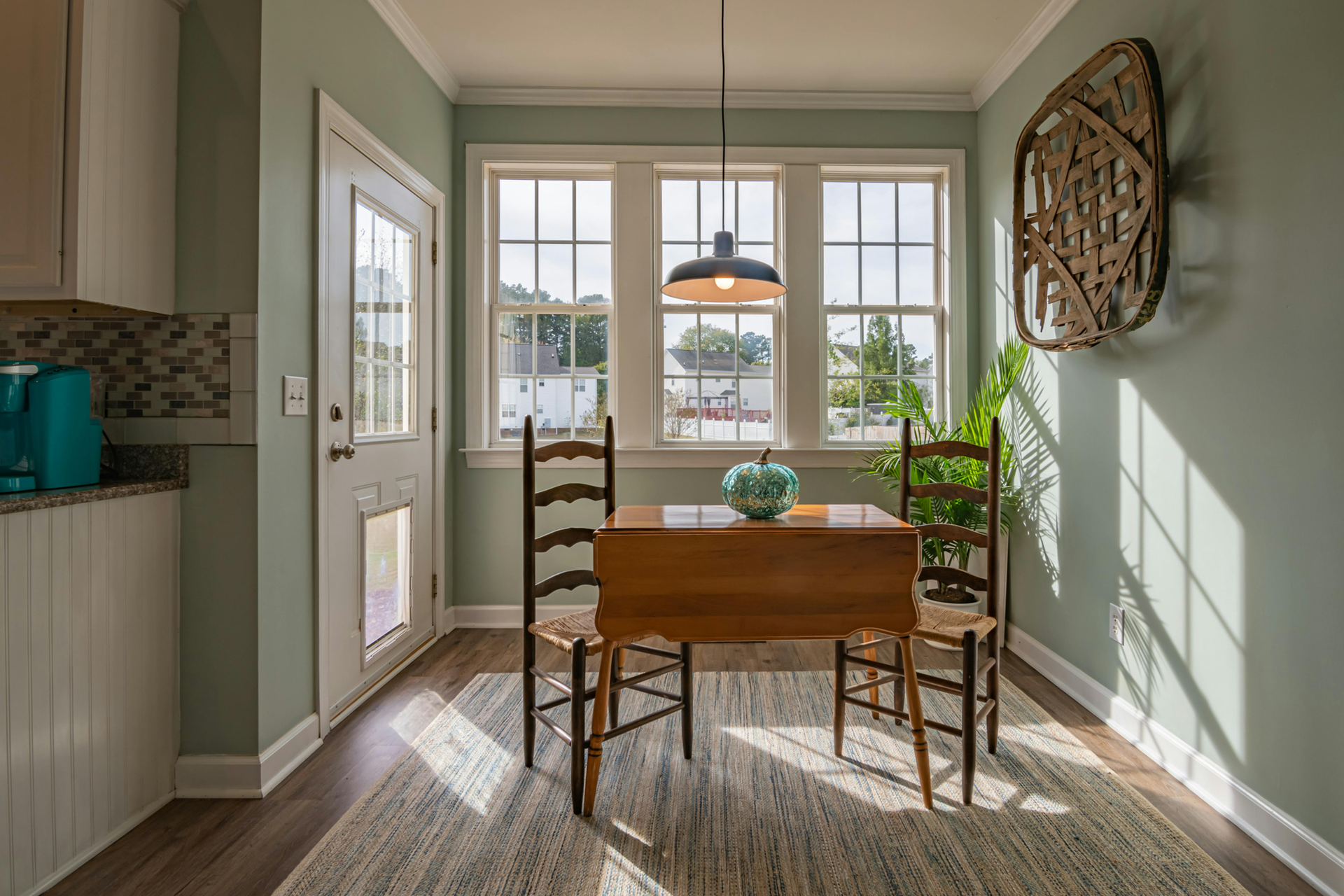 Small kitchen nook with table, chairs, and windows; light green walls, wooden floor and a rug.