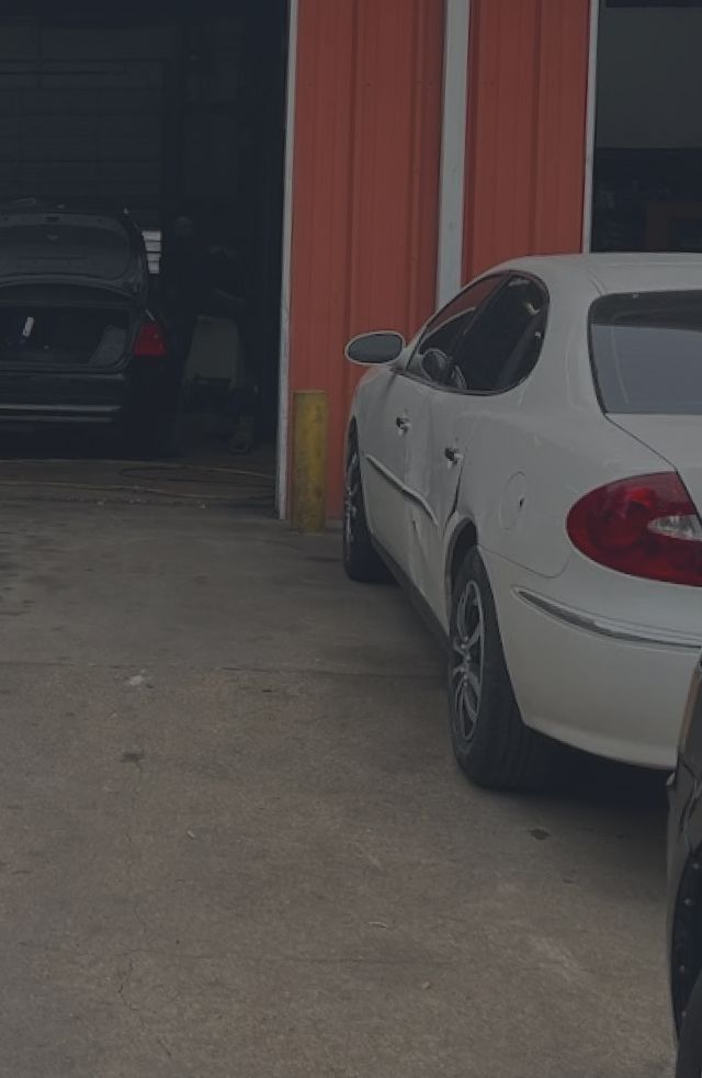 White car parked near a red building; another car inside the garage. | We Fix Cars