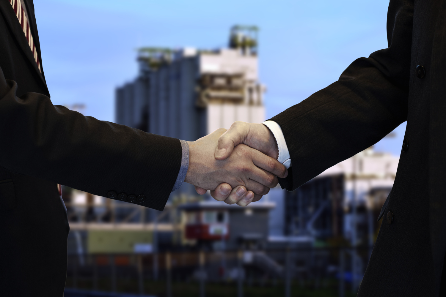 Two businesspeople shaking hands, industrial building in the background, signifying an agreement.