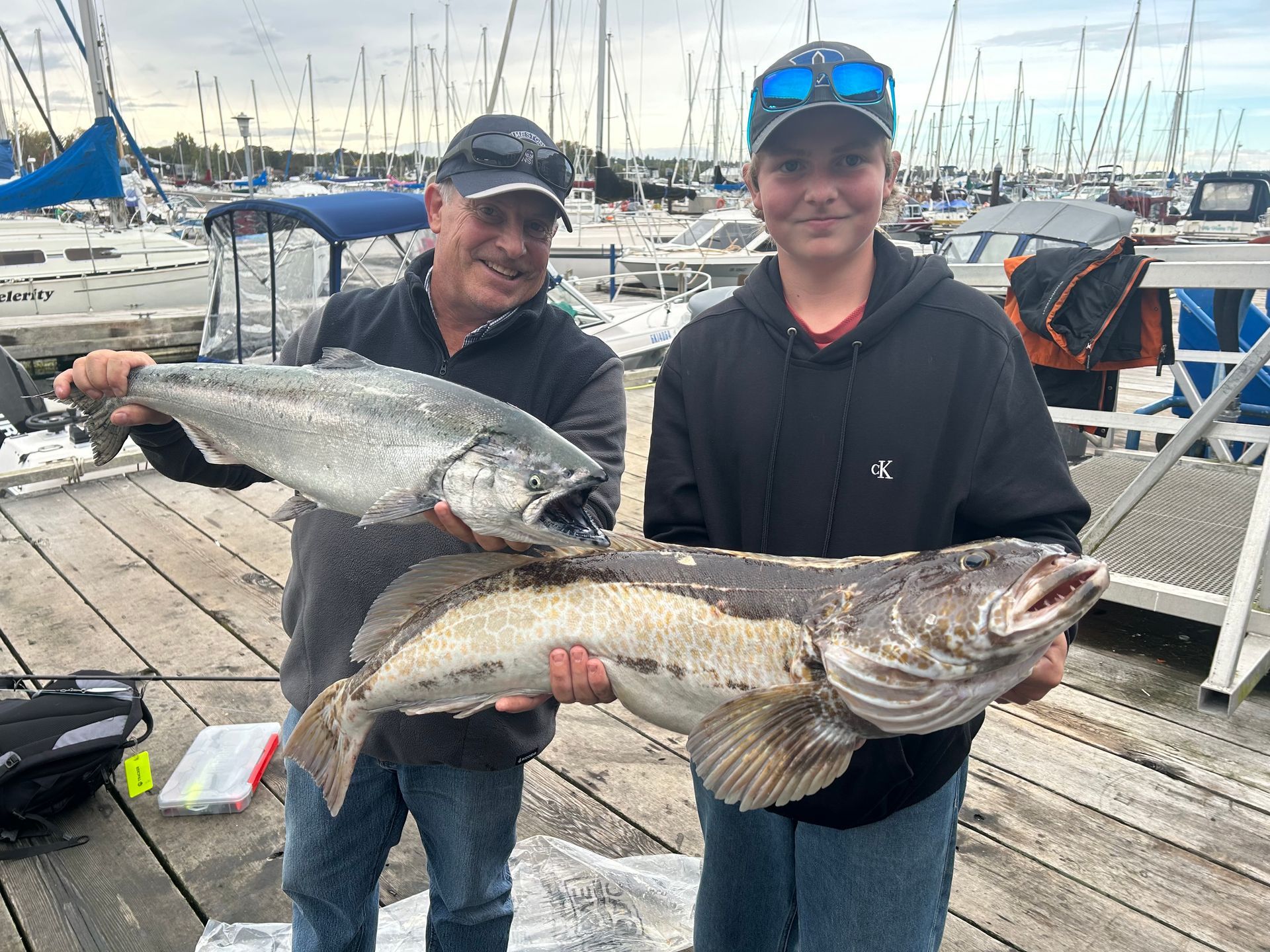 Two people holding large fish at a marina: a silver salmon and a lingcod. Smiling, cloudy day.