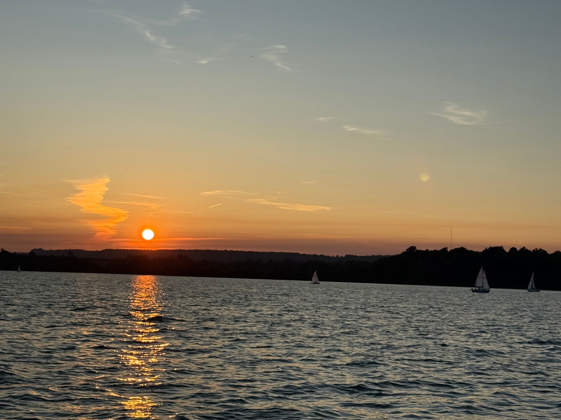 Sunset over water, sailboats on the horizon, orange and yellow sky, sun reflecting on water.