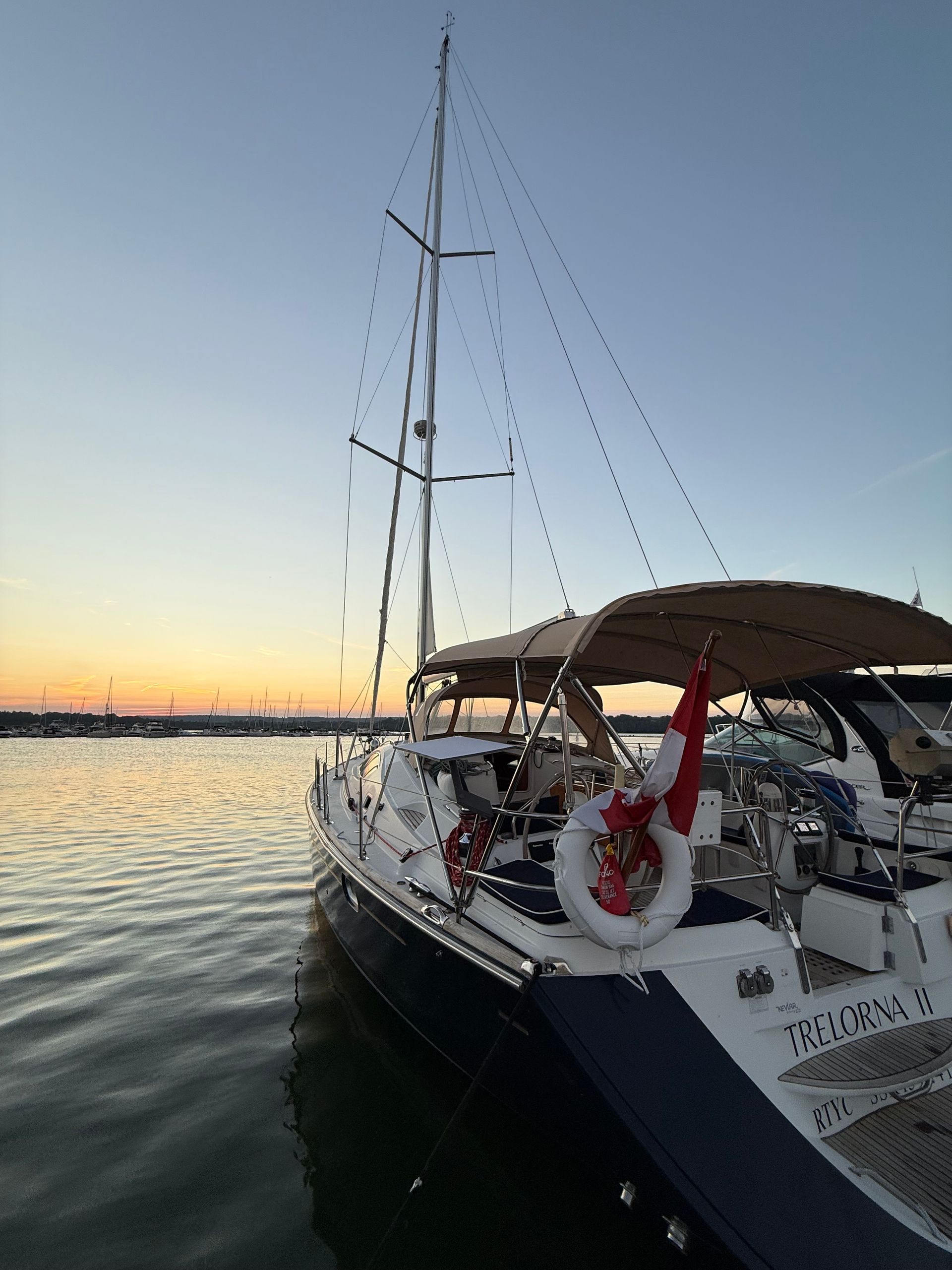 Sailboat at dock, dusk sky. Beige awning, red and white life preserver. Calm water, city skyline.