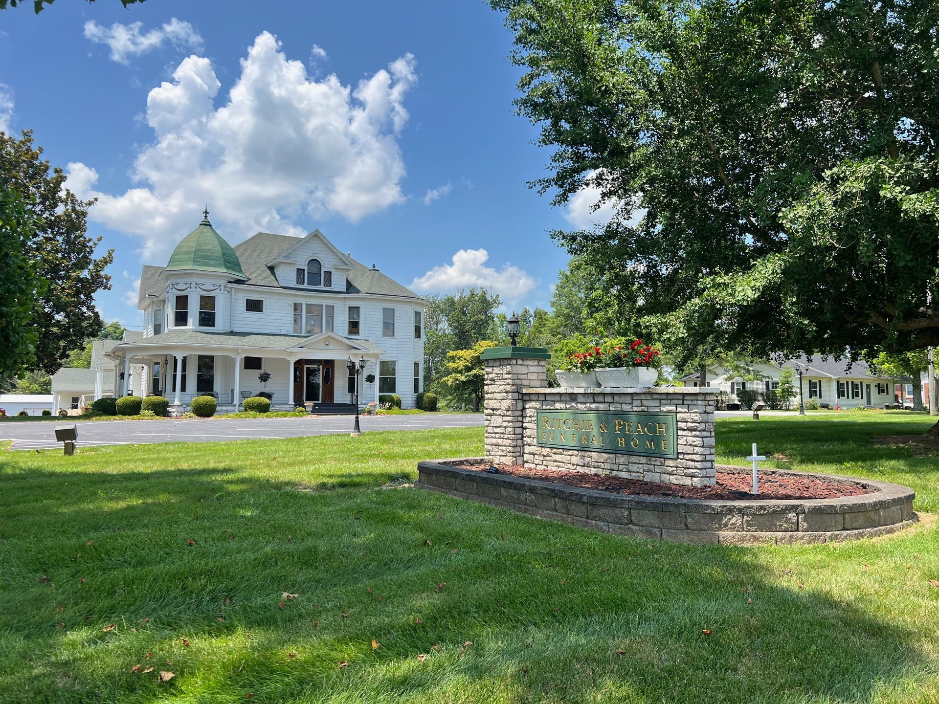 White mansion with green roof and brick fountain on a sunny lawn under blue sky