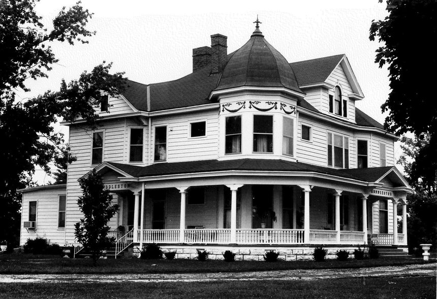 White Victorian house with wrap-around porch and turret, trees in the background.