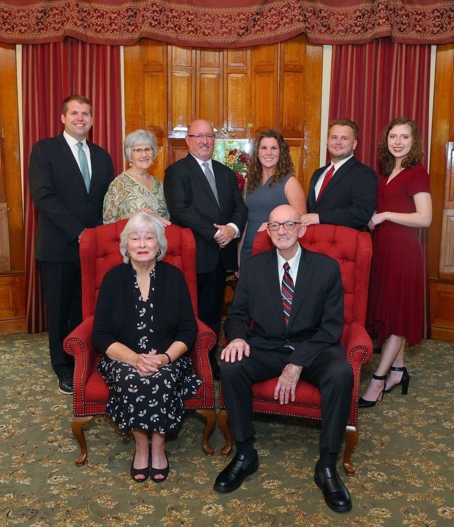 Family group portrait, posed indoors. Two seated, others standing behind. Formal attire, red curtains.