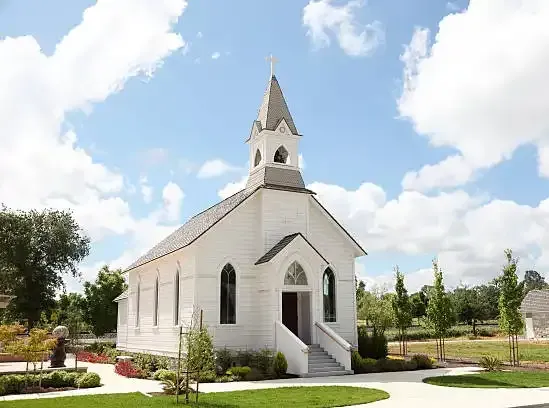 Small white chapel with steeple and arched entrance in a landscaped garden under a blue sky