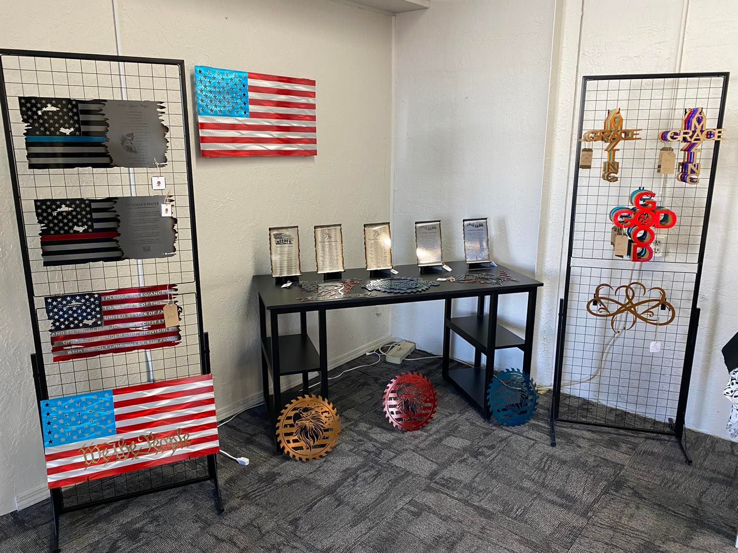 Display of metal art: American flags, crosses, and other pieces on black stands and table in a room with gray carpet.