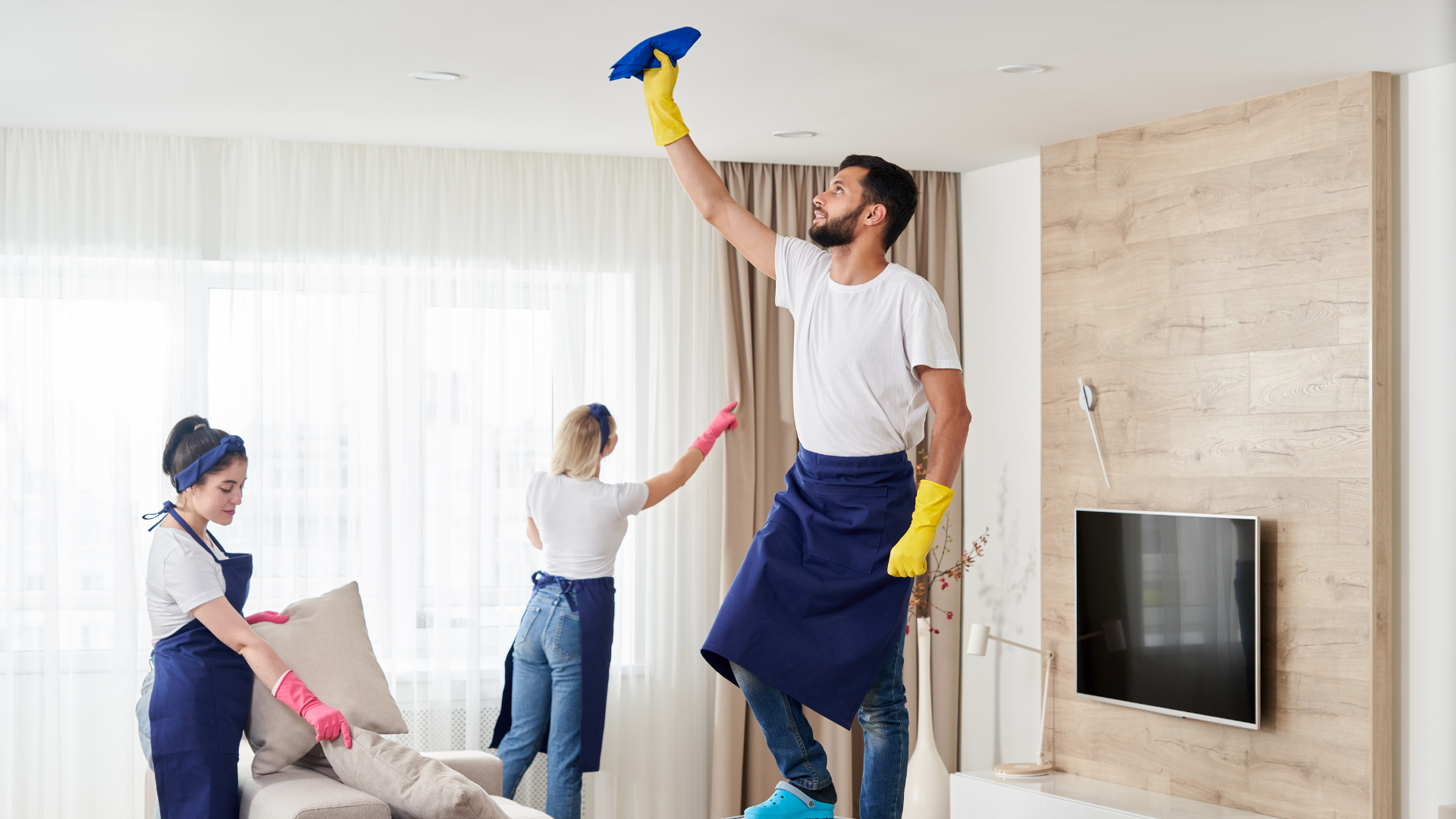 Three people cleaning a living room: one on a step stool dusting the ceiling, two dusting a couch.