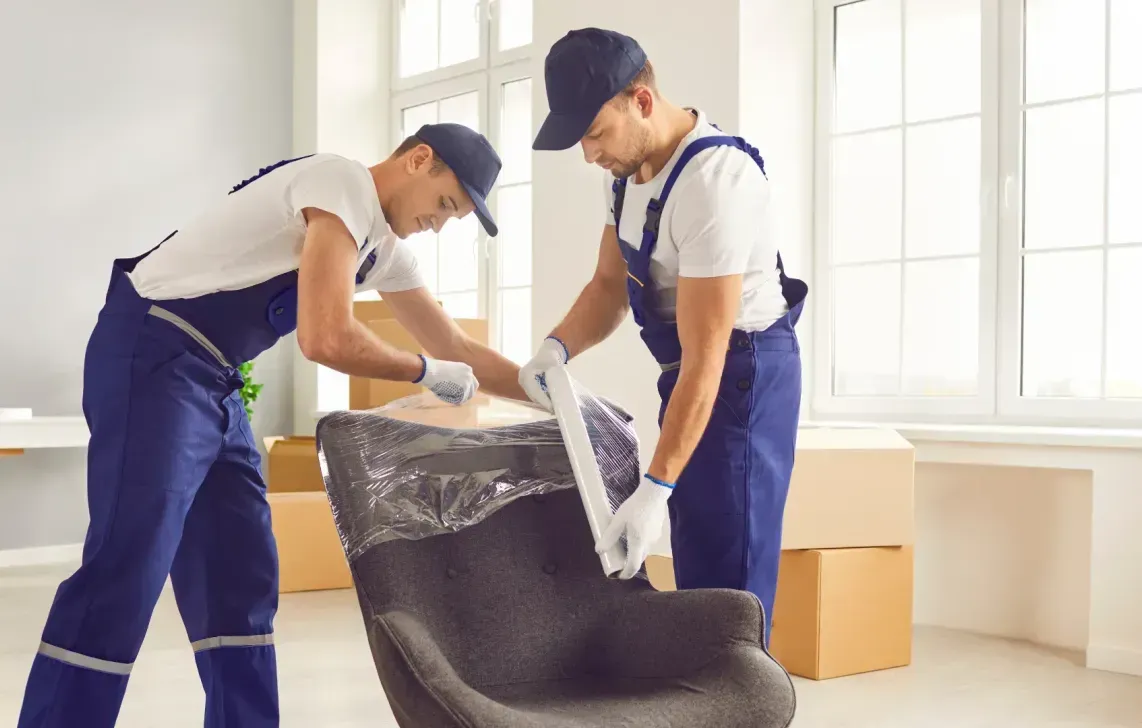 Two movers in blue coveralls wrapping a dark chair with plastic wrap in a room with cardboard boxes.