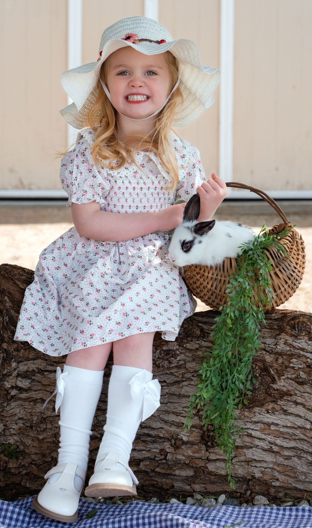 A little girl is sitting on a log holding a basket and a rabbit.