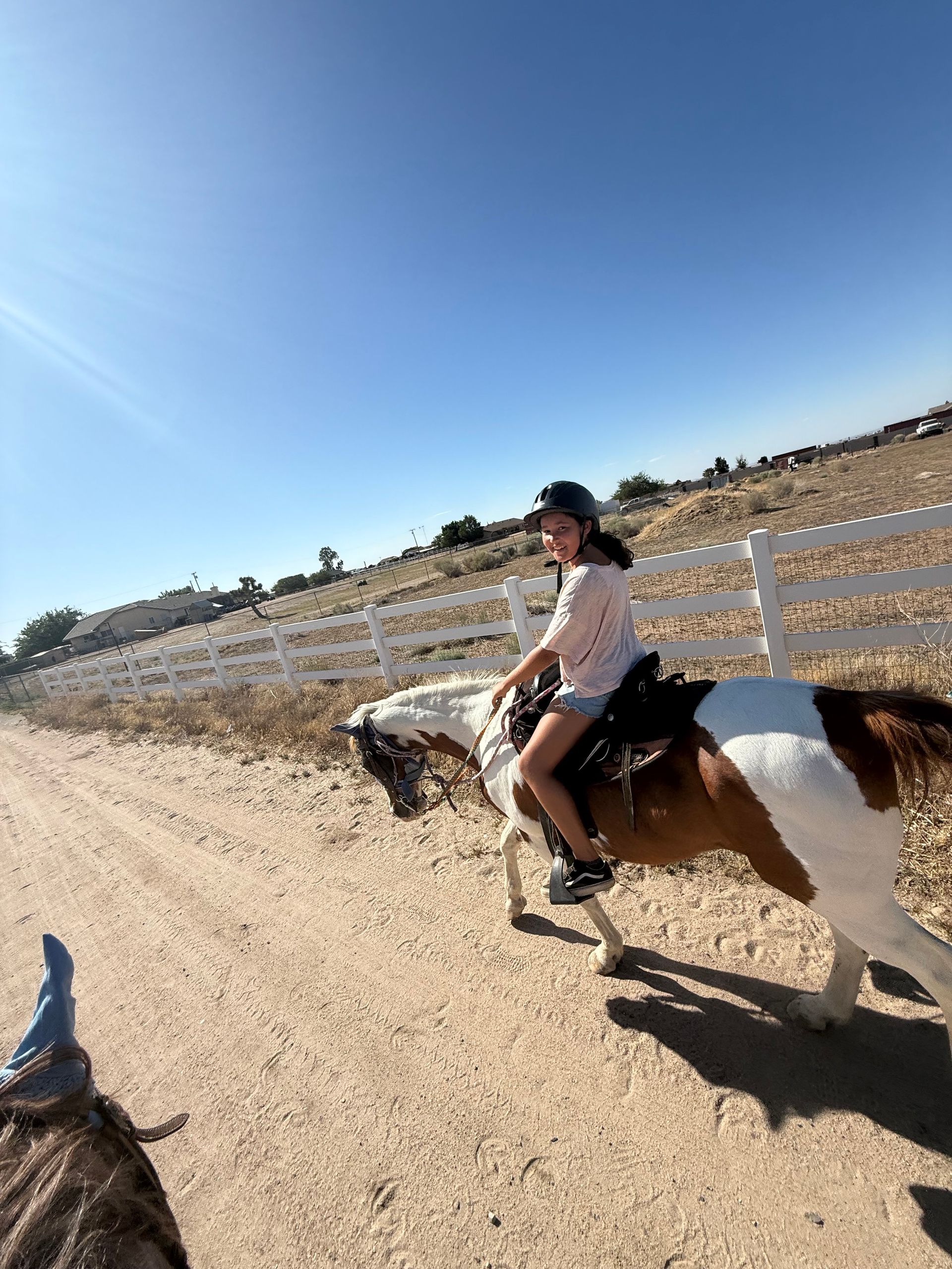 Girl riding a paint horse on a dirt path with a white fence and blue sky in the background.
