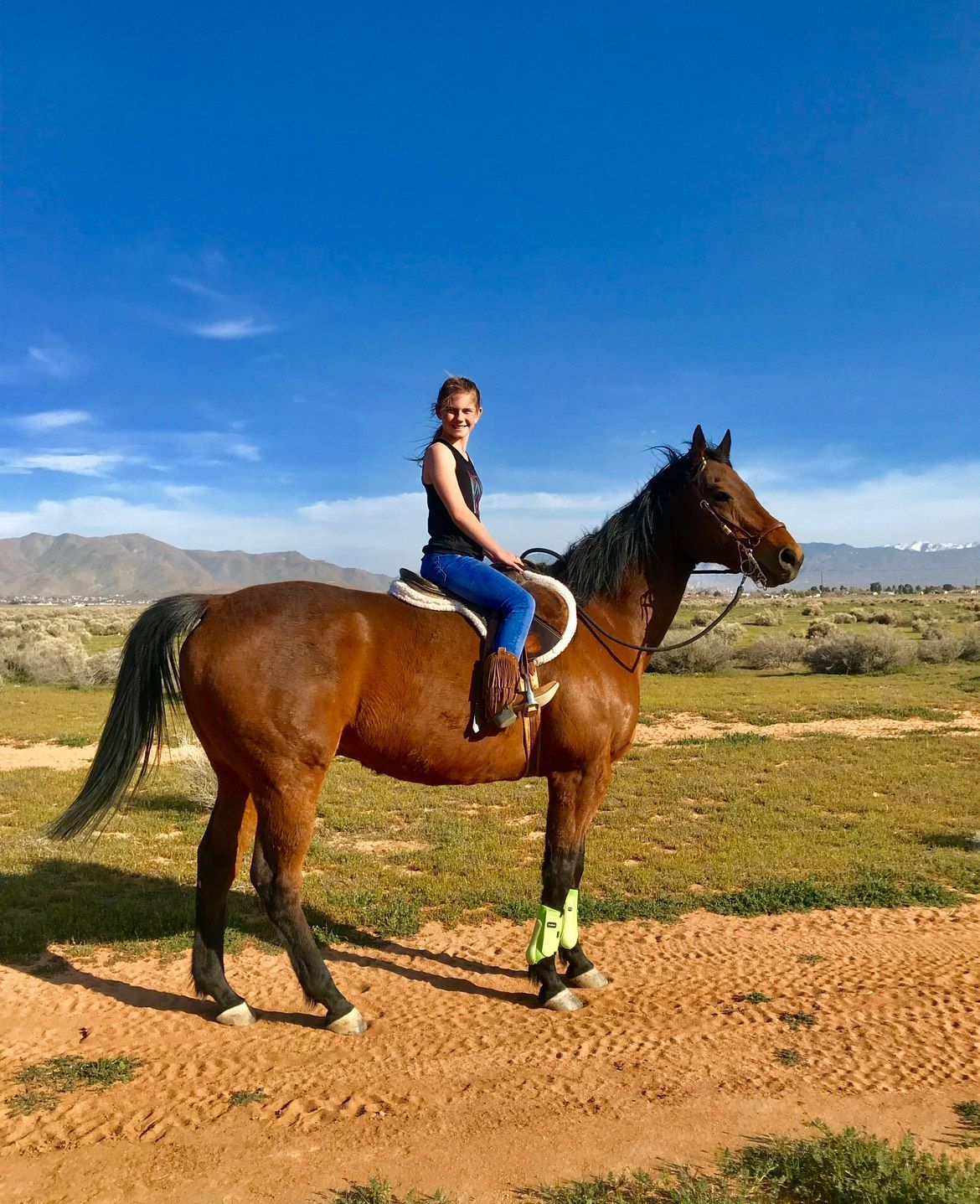 Young person riding a brown horse on a dirt path in a sunny, desert-like landscape.