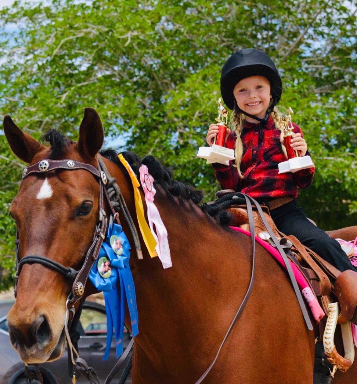 Girl in helmet on horse, holding trophies, smiling. Horse has ribbons.